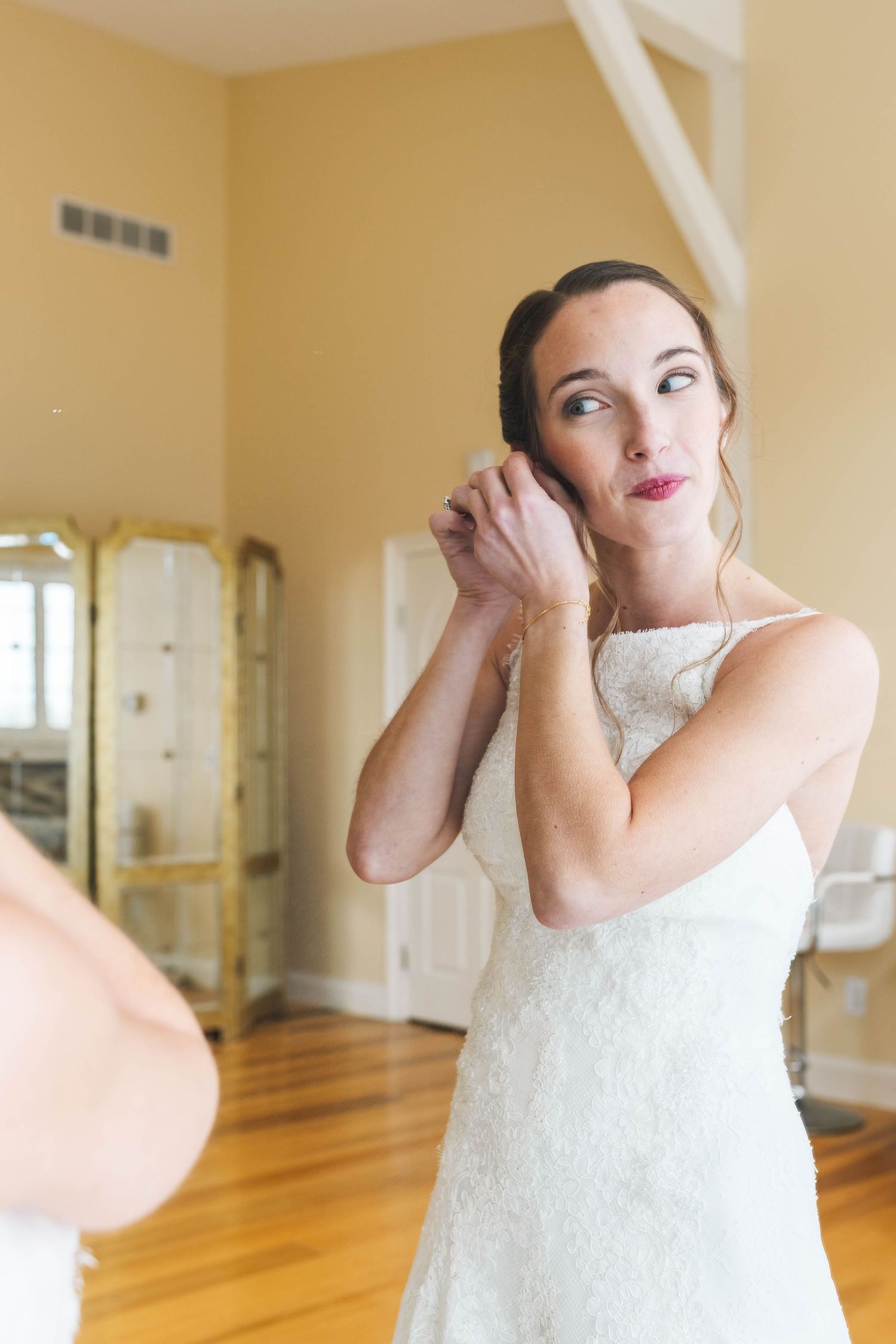 Bride in lace wedding dress putting on earrings while getting ready in a bright room.
