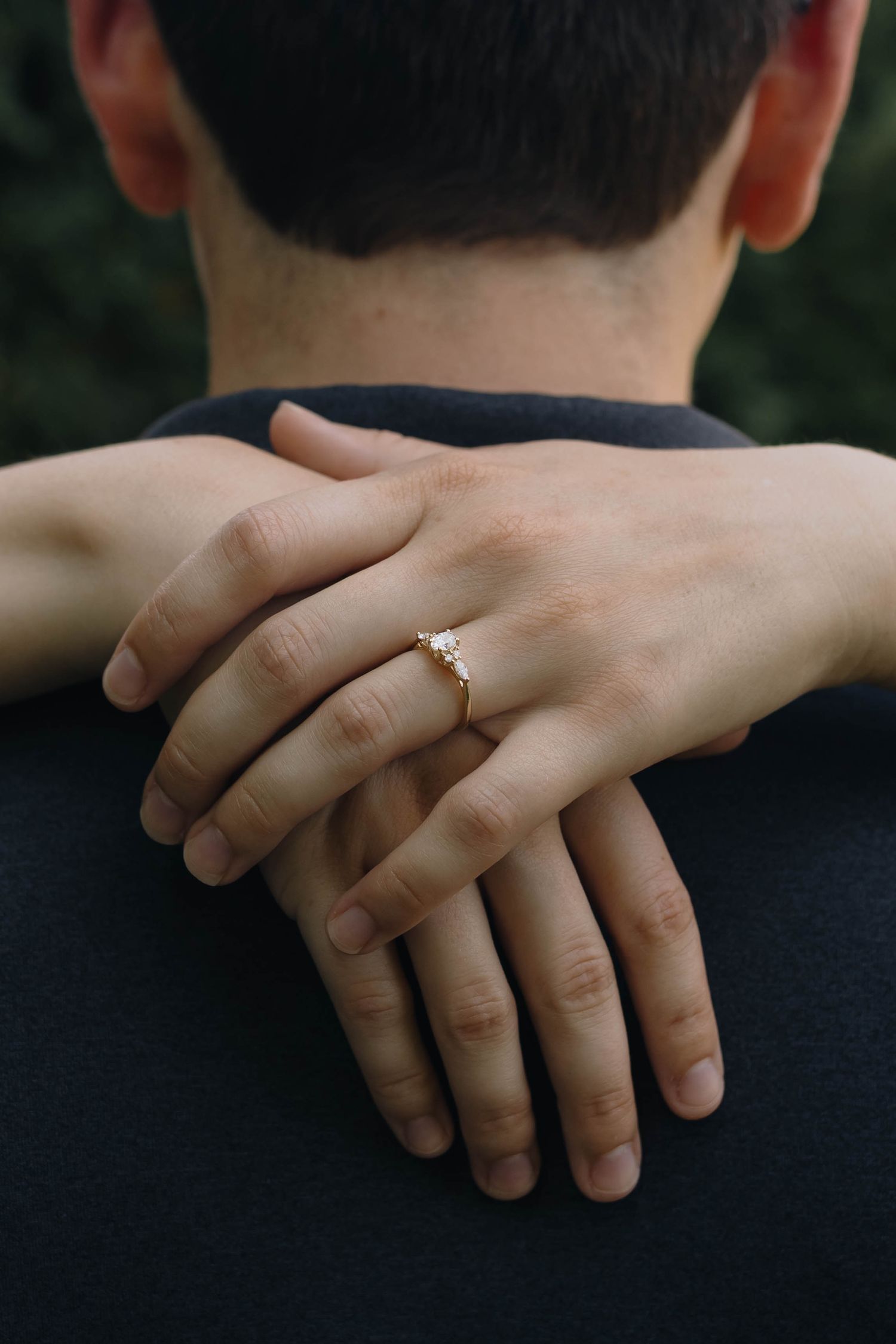 Close-up of a woman's hands with a diamond engagement ring resting on a man's shoulders from behind.