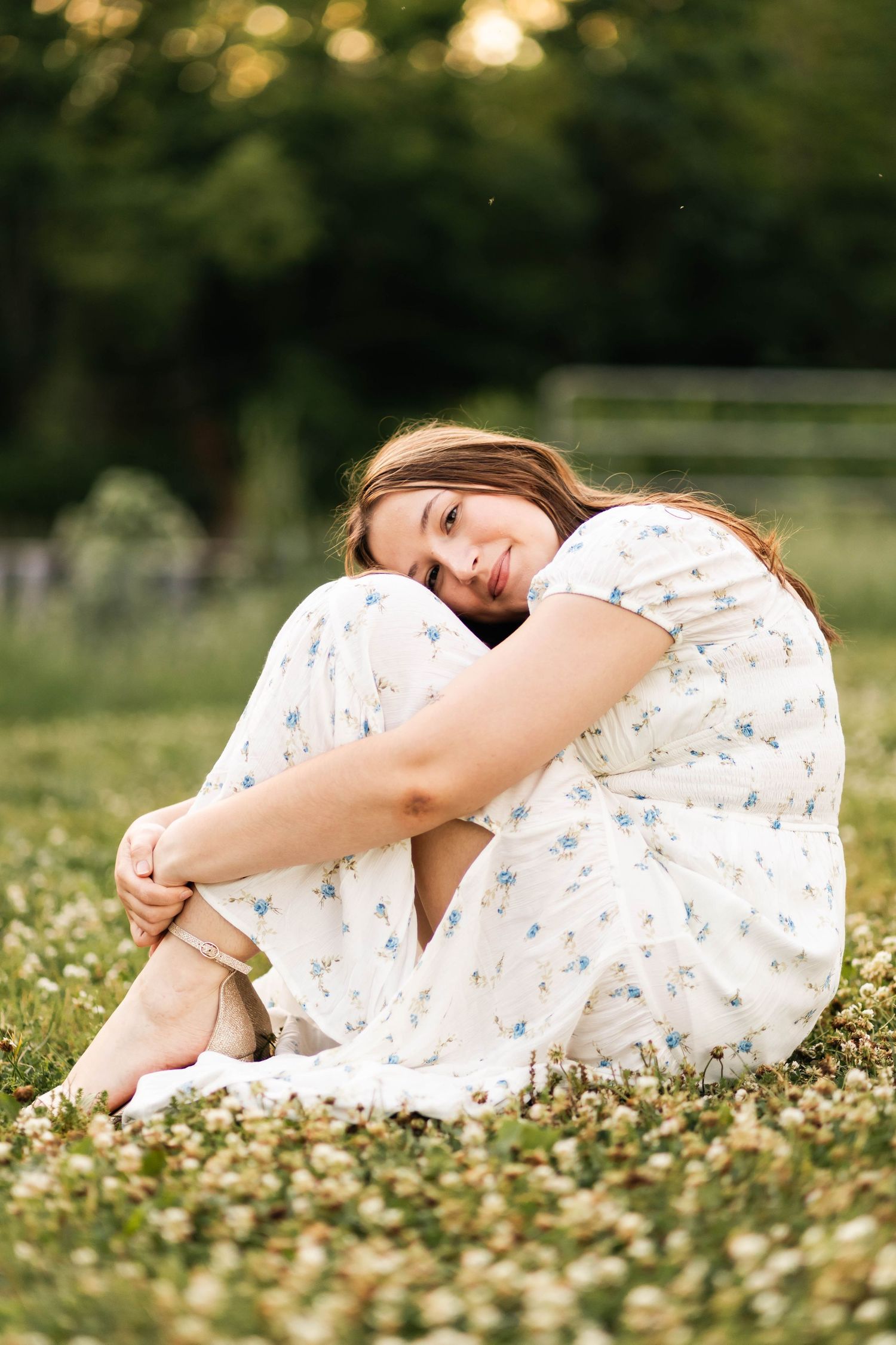 Boyertown Senior in white floral dress sitting in a field of wildflowers, hugging her knees and smiling softly in Harleysville, PA.