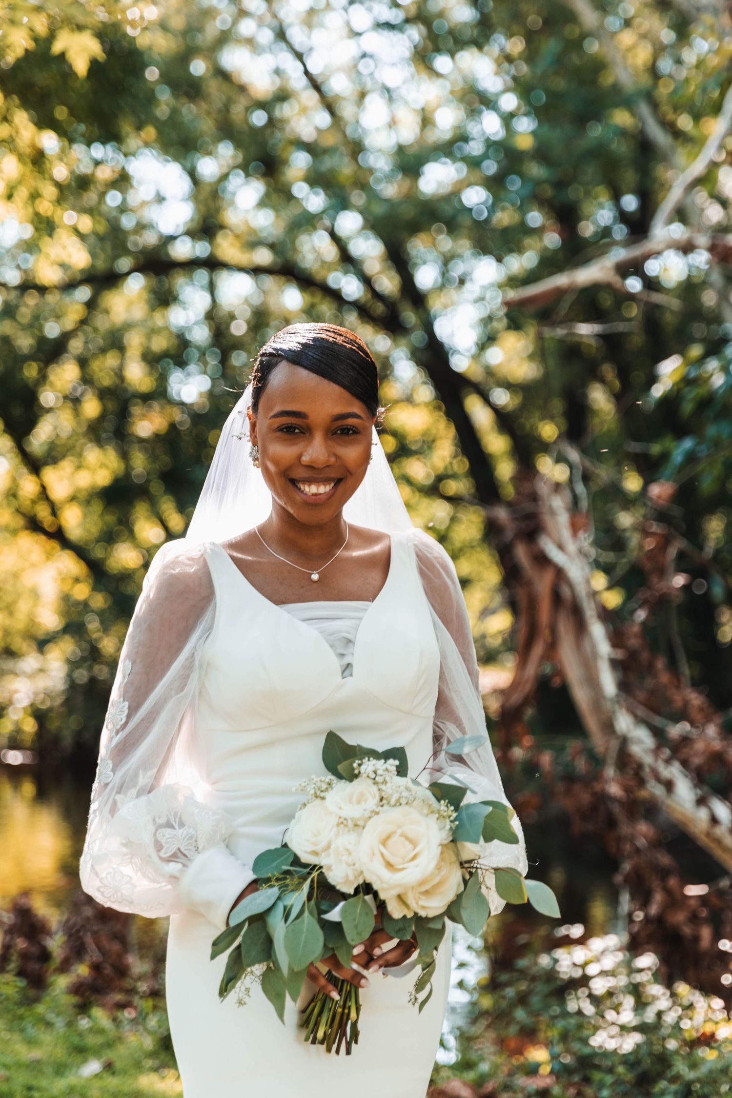Smiling Black bride in white gown and veil holding white rose bouquet in garden setting.