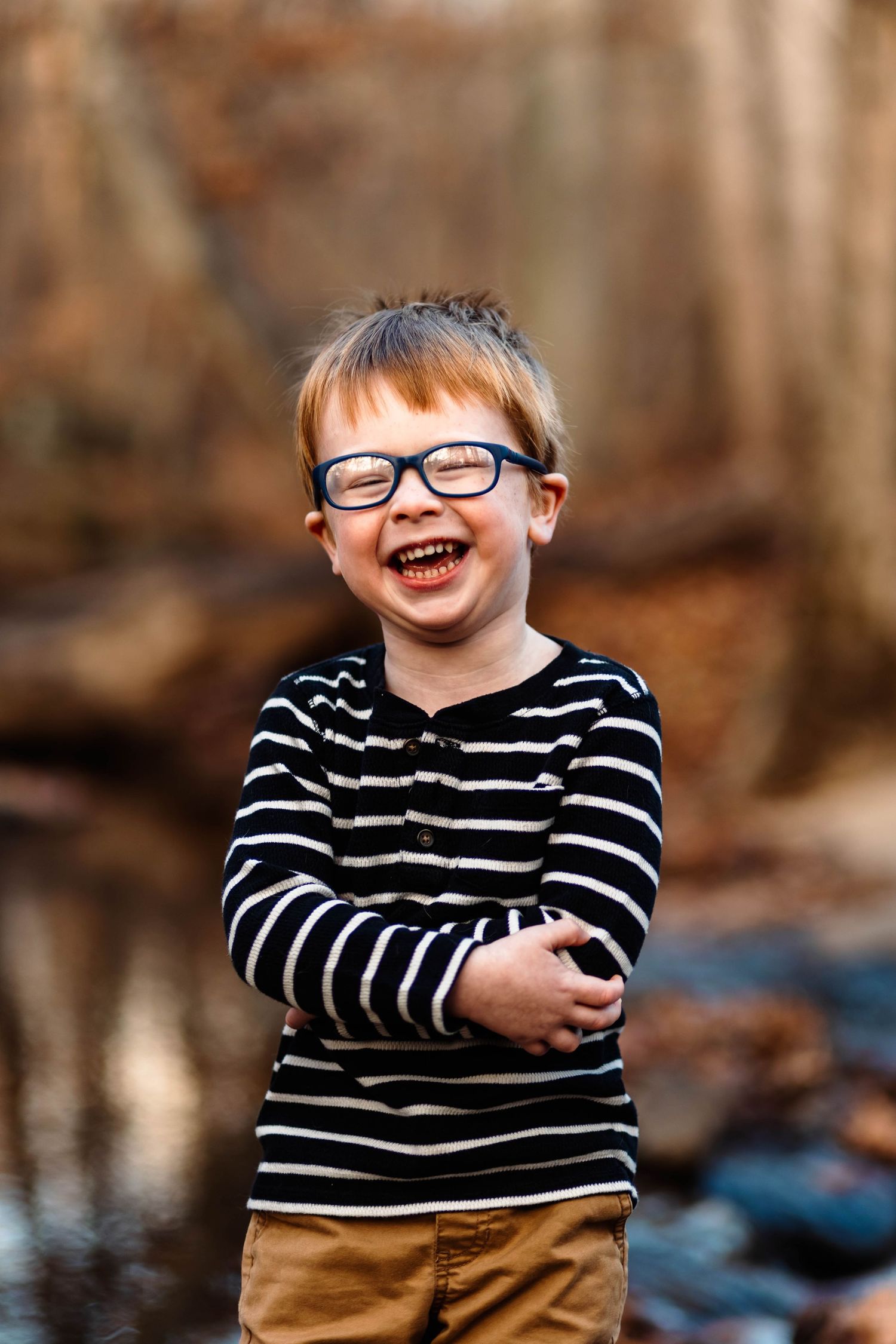 An adorable laughing boy wearing glasses and a striped shirt stands with arms crossed near a creek in Pottstown, PA.