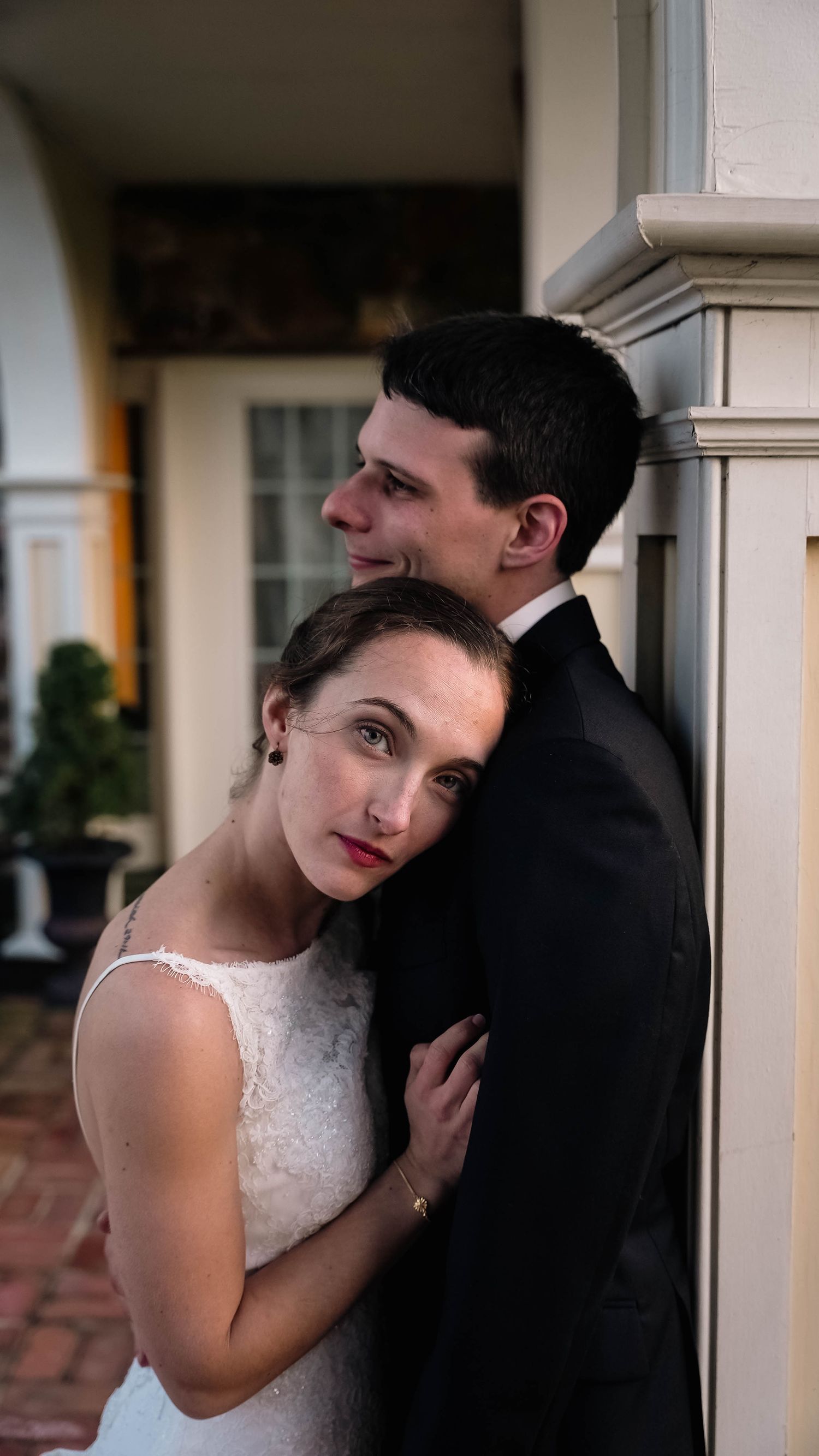 Bride in white lace dress leans against groom in black tuxedo during romantic wedding portrait near elegant column in Chester County.