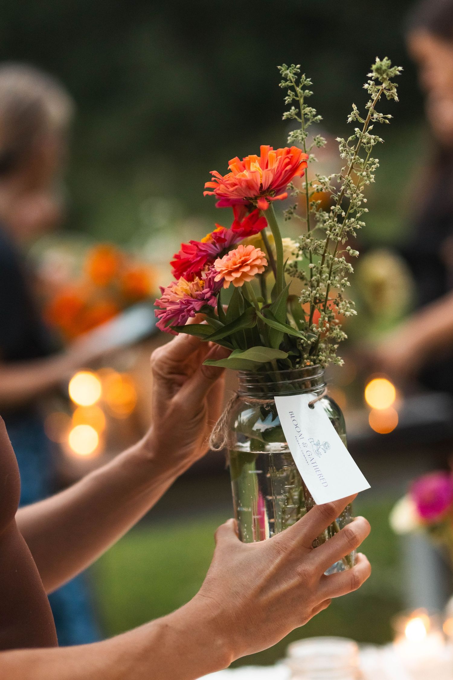 Hands holding a mason jar filled with vibrant orange and red zinnias at a candlelit outdoor gathering.