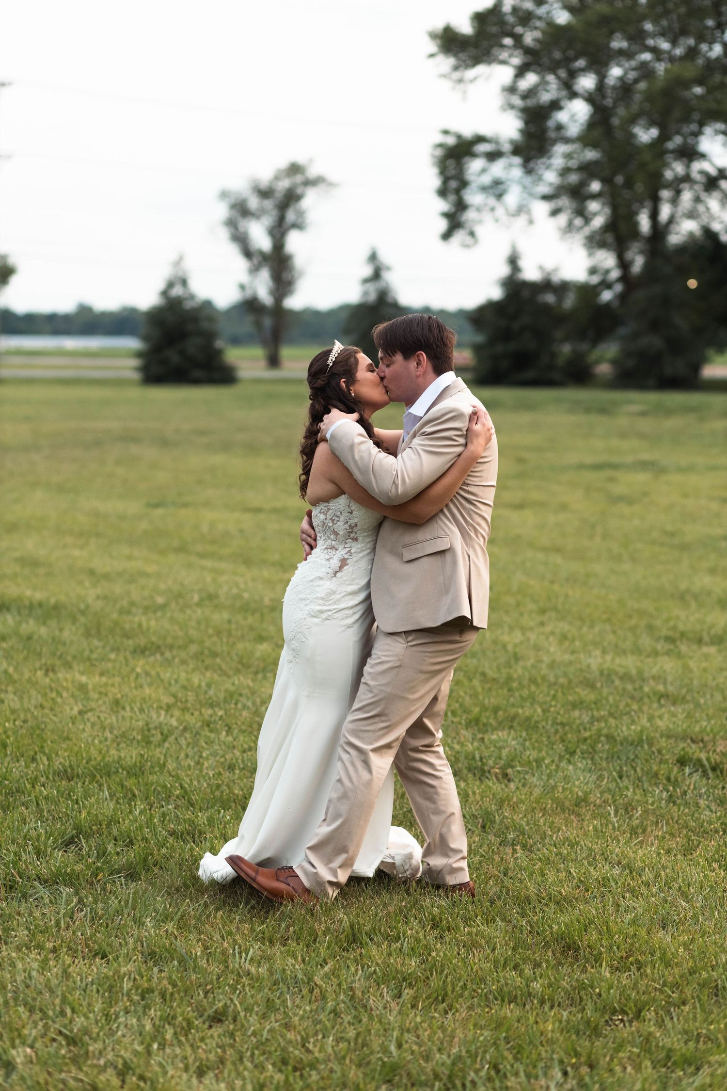 Bride and groom share a romantic kiss while embracing in a wide open green field on their wedding day.