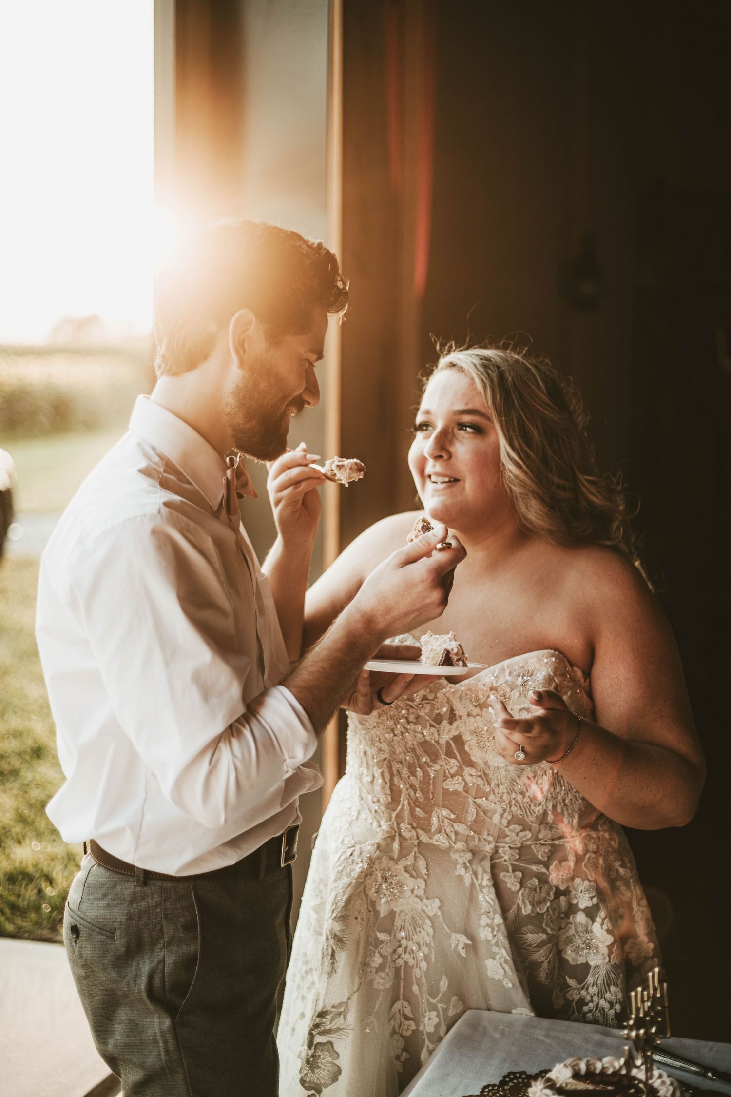 Bride and groom share wedding cake bathed in golden sunset light during outdoor reception.