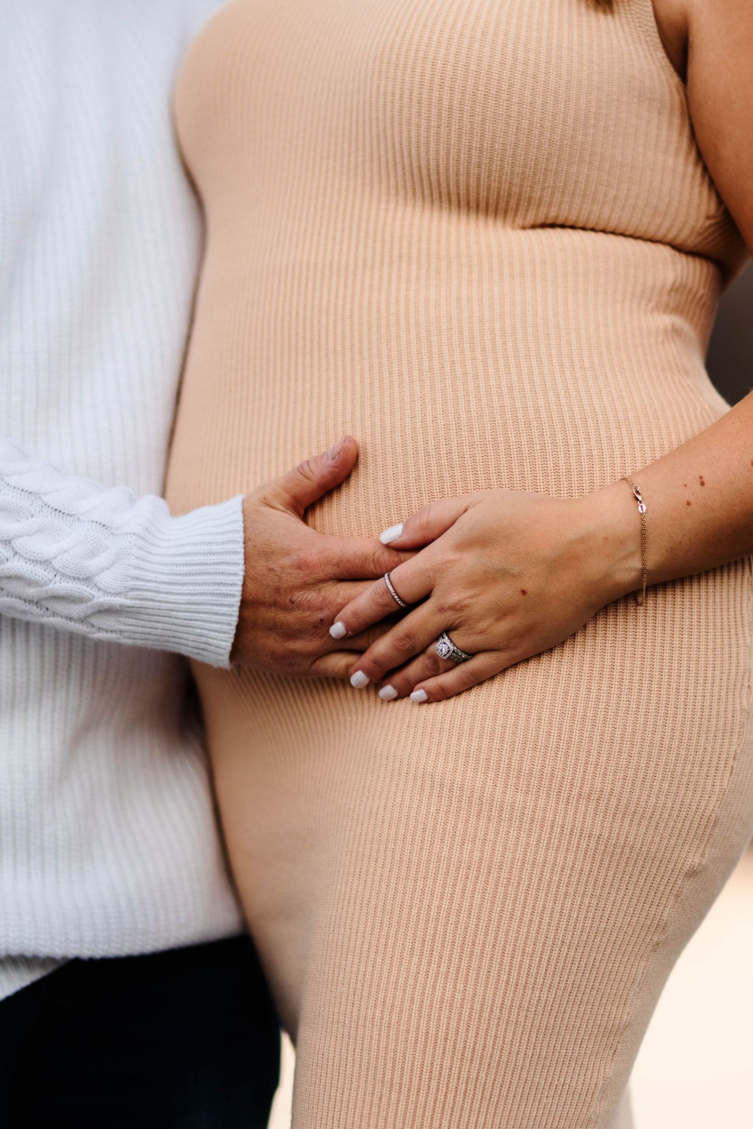 Maternity session of expecting mother in beige dress with partner's hands resting on baby bump at Green Lane Park.