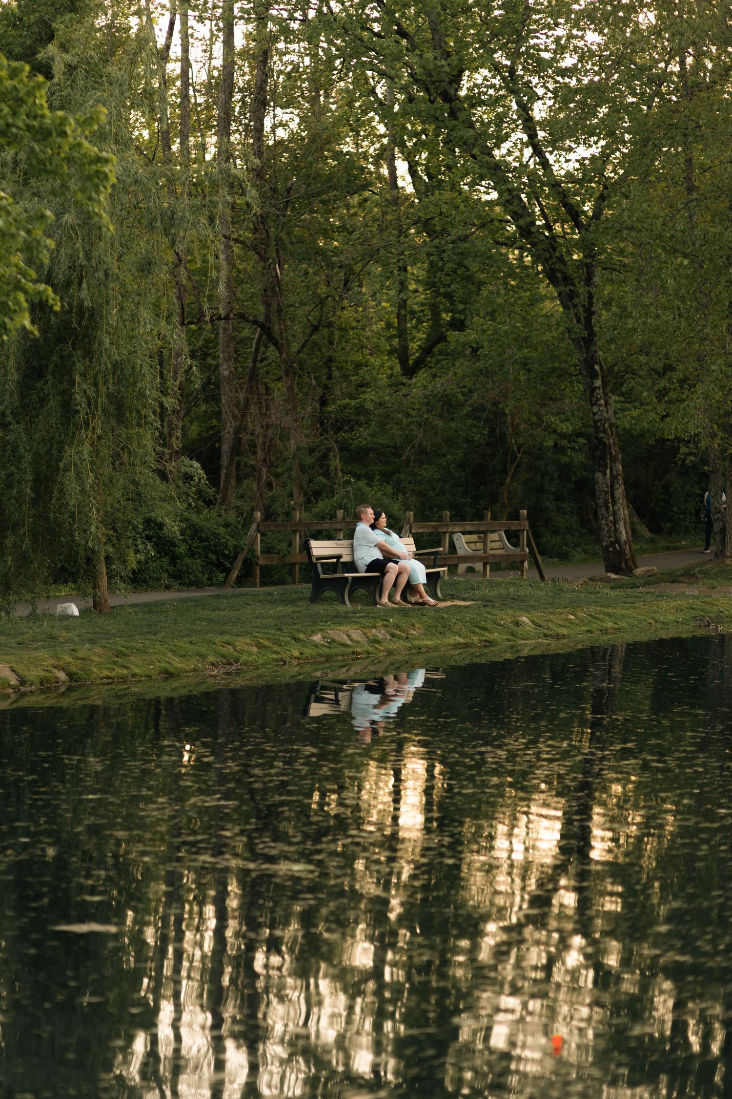 Expecting couple sitting on a bench by a tranquil pond surrounded by lush green trees in Philadelphia, PA.