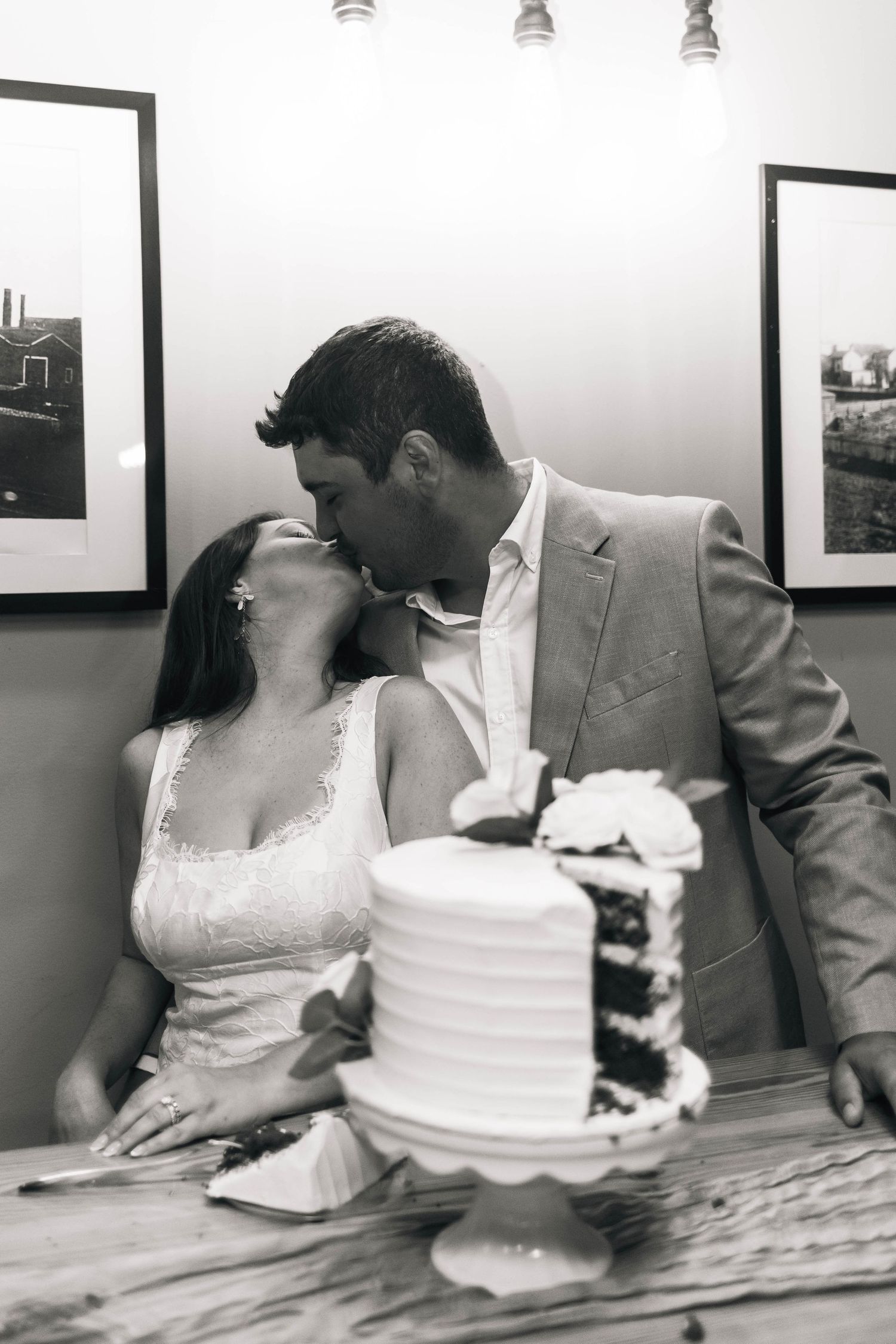 Black and white photo of bride and groom sharing a kiss behind their white wedding cake at reception.