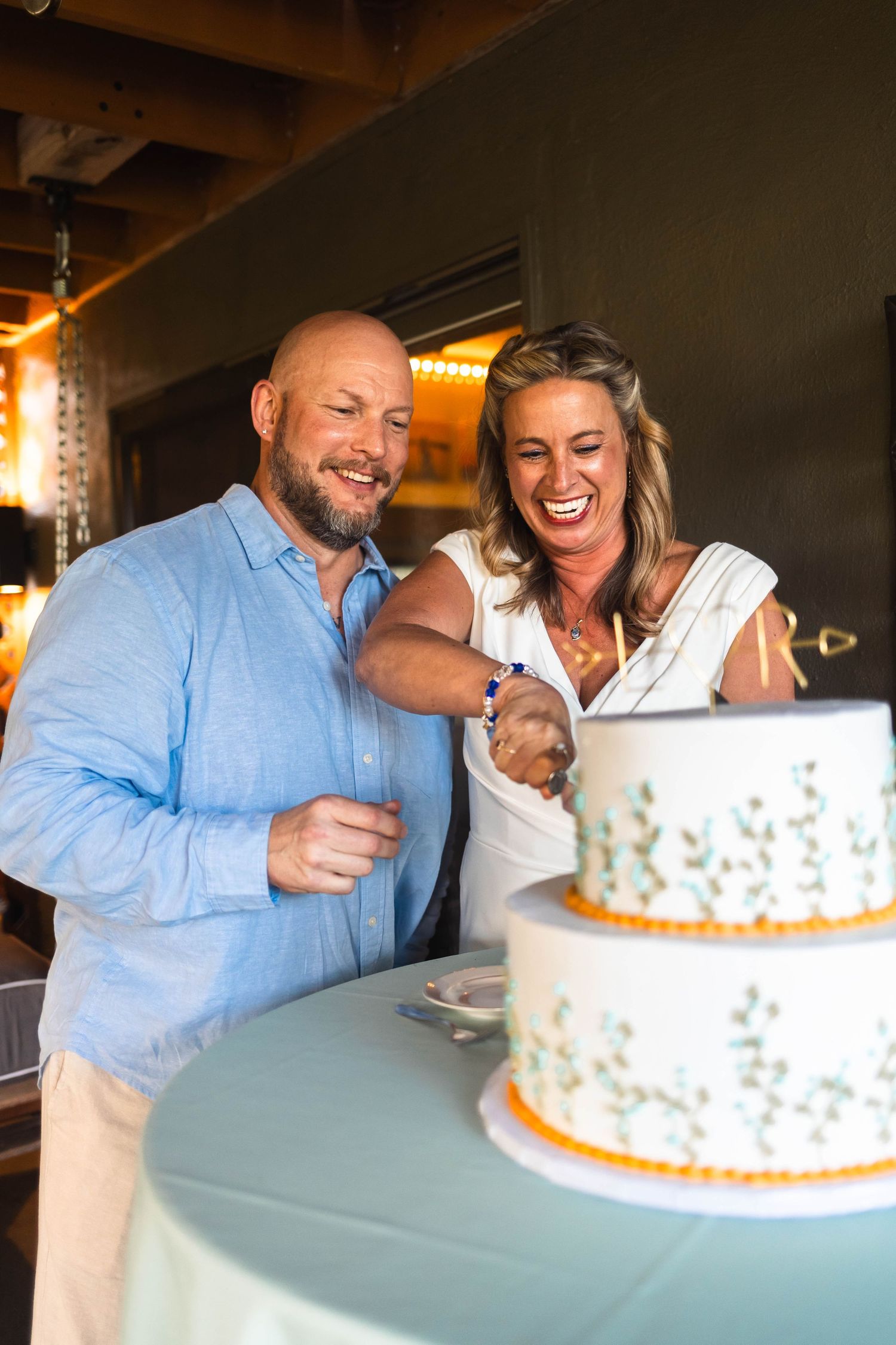 Laughing couple cutting their two-tier white wedding cake decorated with floral designs indoors.