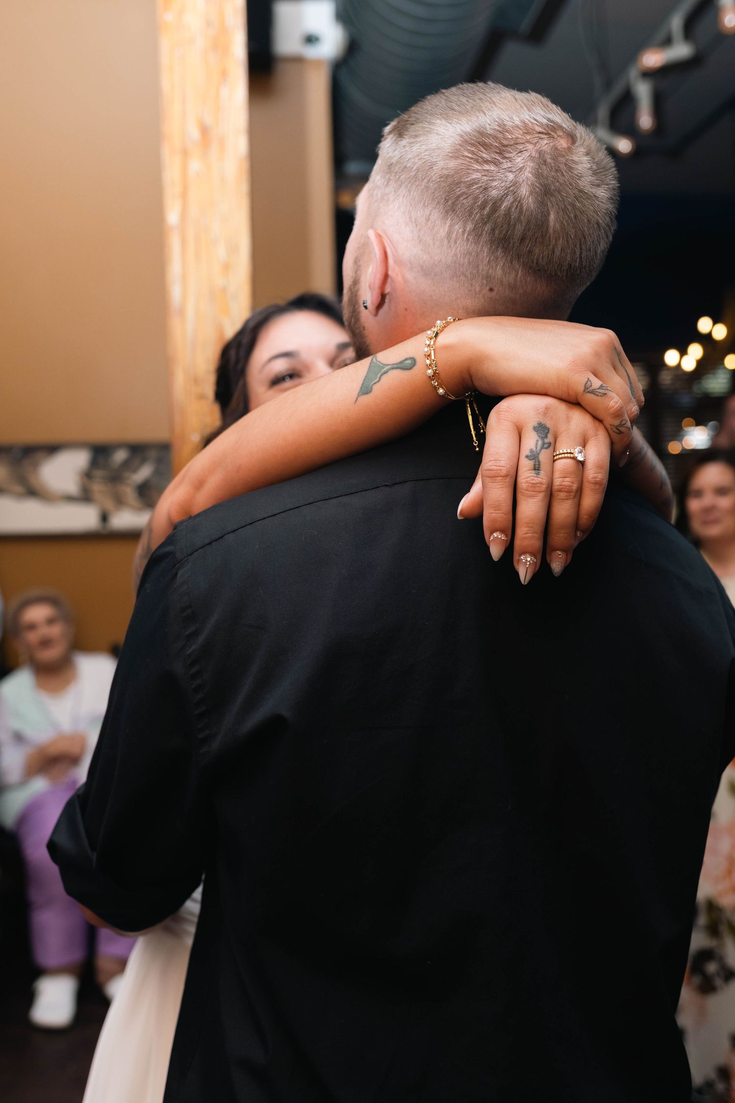 Bride with tattoos embracing groom during first dance, showing wedding ring and gold bracelet details.