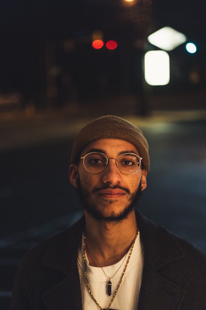 Young man with round glasses and beanie hat standing on city street at night with bokeh lights.