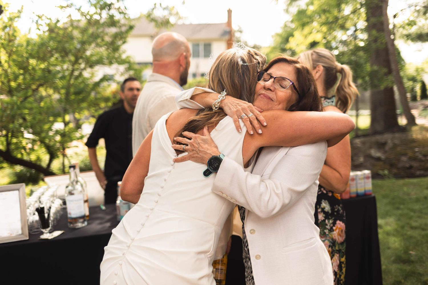 Bride in white dress receiving warm embraces from emotional guests at outdoor wedding reception.