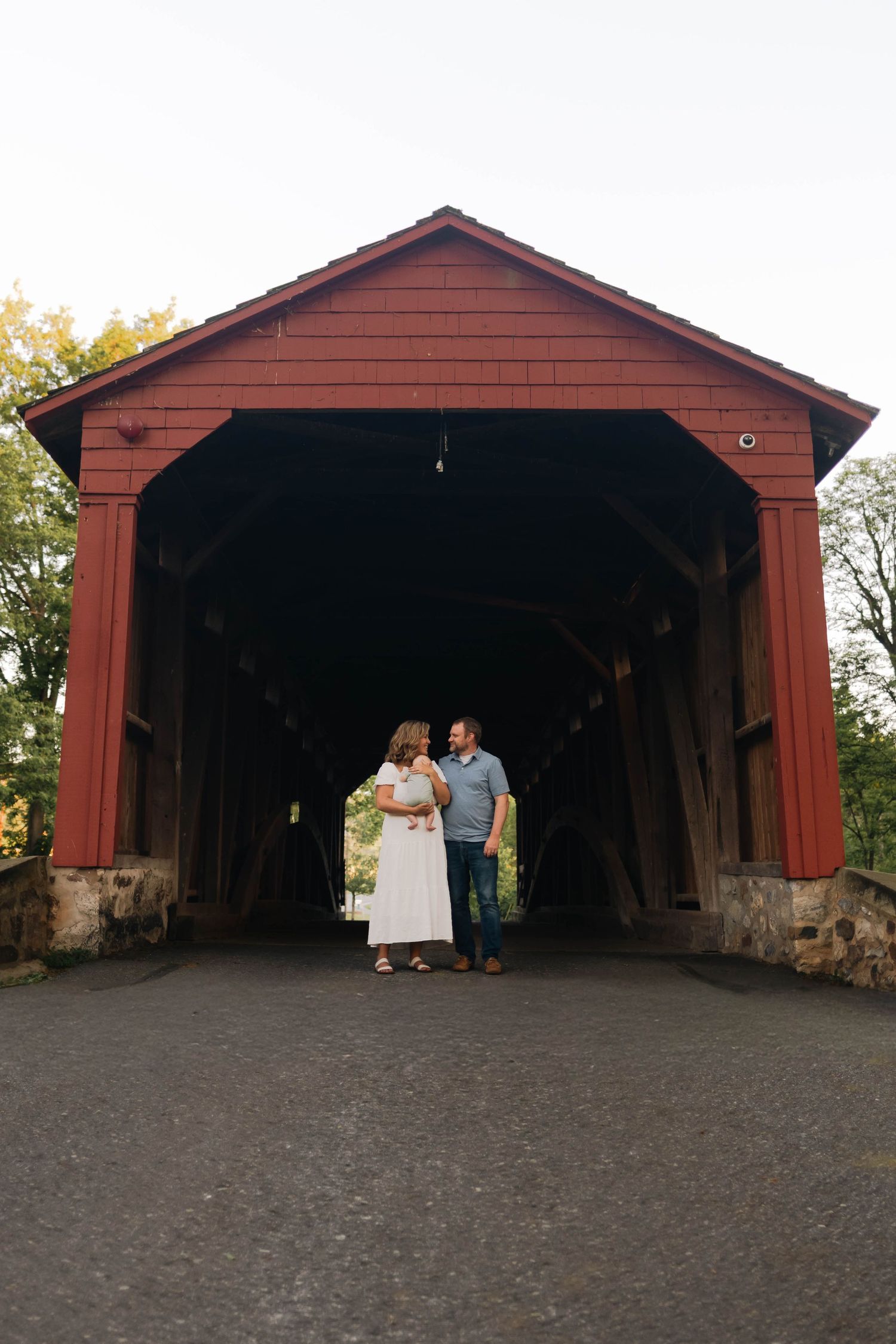 Couple standing together at the entrance of a historic red covered bridge on a cloudy day in Elverson, PA.
