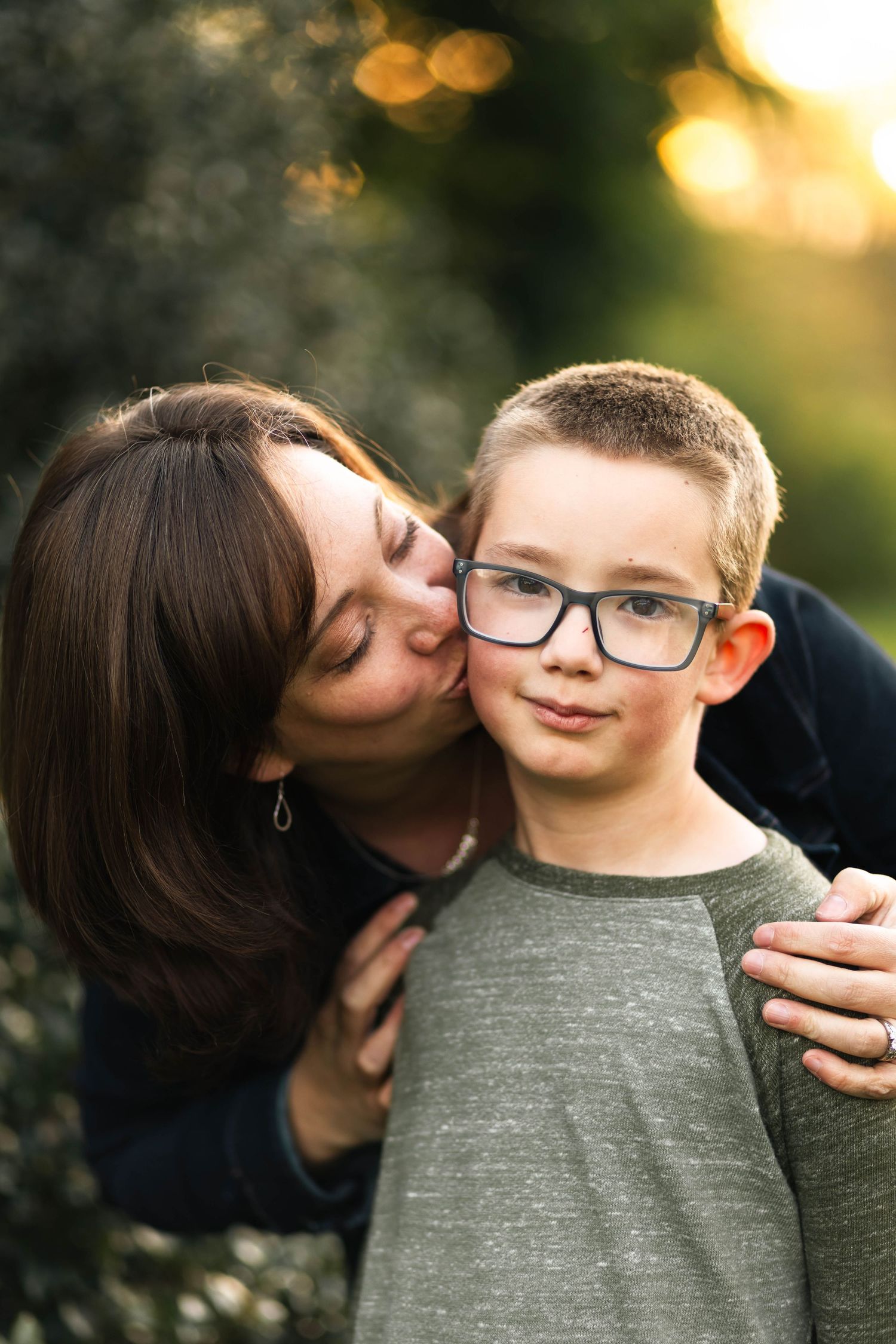 A dark-haired mother lovingly kisses her young son wearing glasses on the cheek in a sunlit outdoor setting in Elverson, PA.