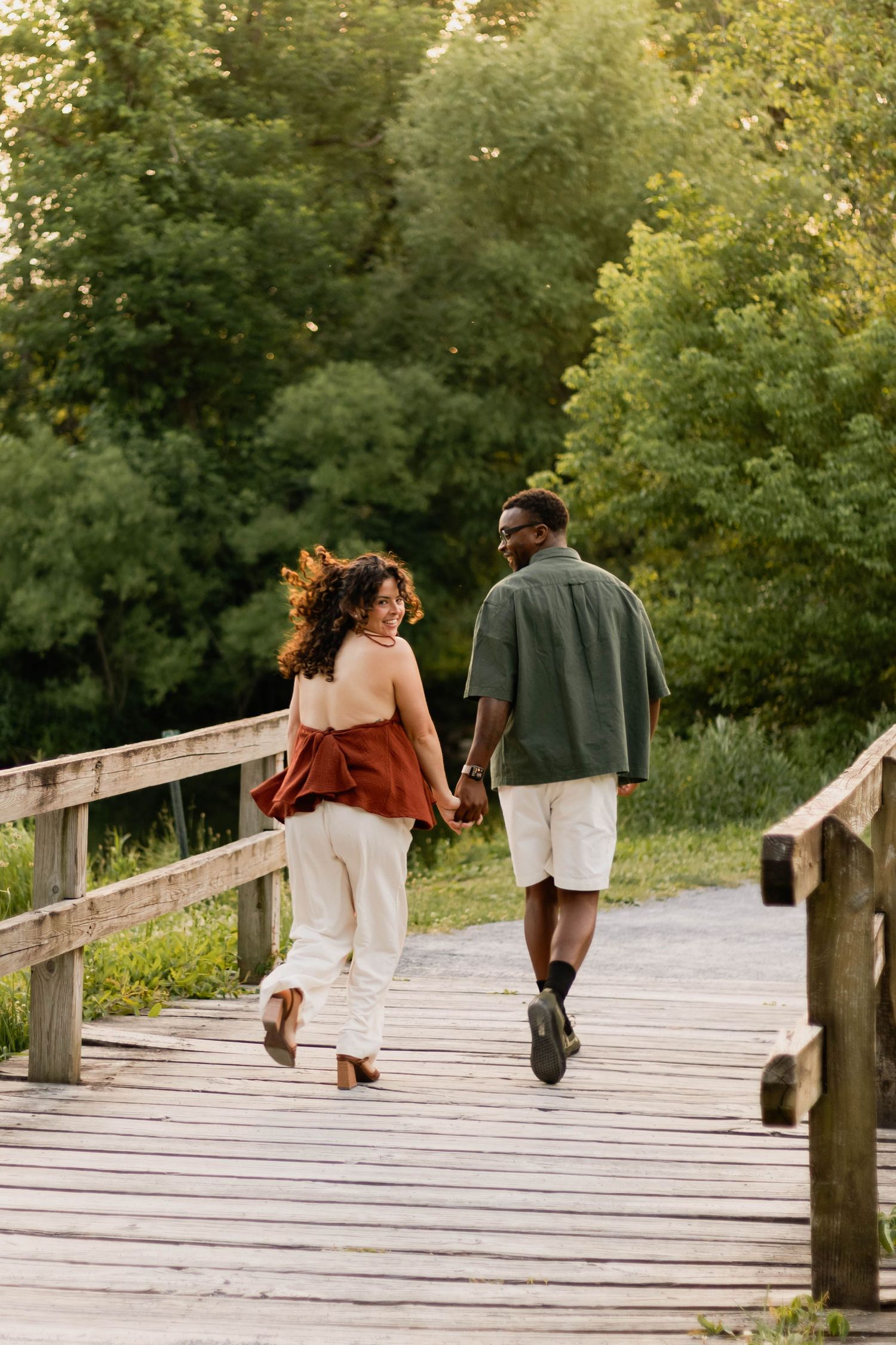 An engaged couple walks hand in hand across a wooden bridge surrounded by lush green trees at sunset at Grings Mill in Reading, PA.