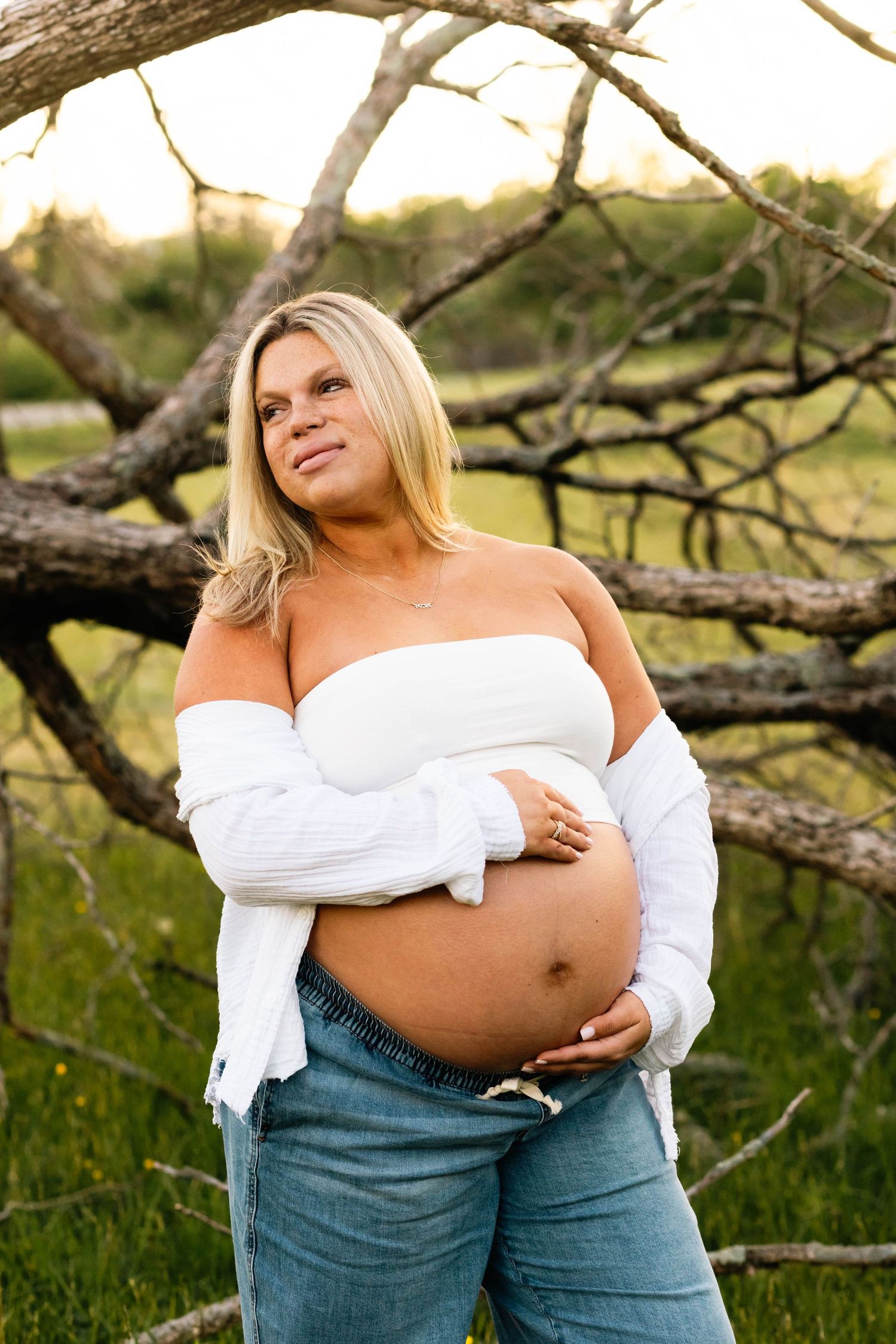 Pregnant blonde woman in white crop top and jeans cradling baby bump outdoors at golden hour.