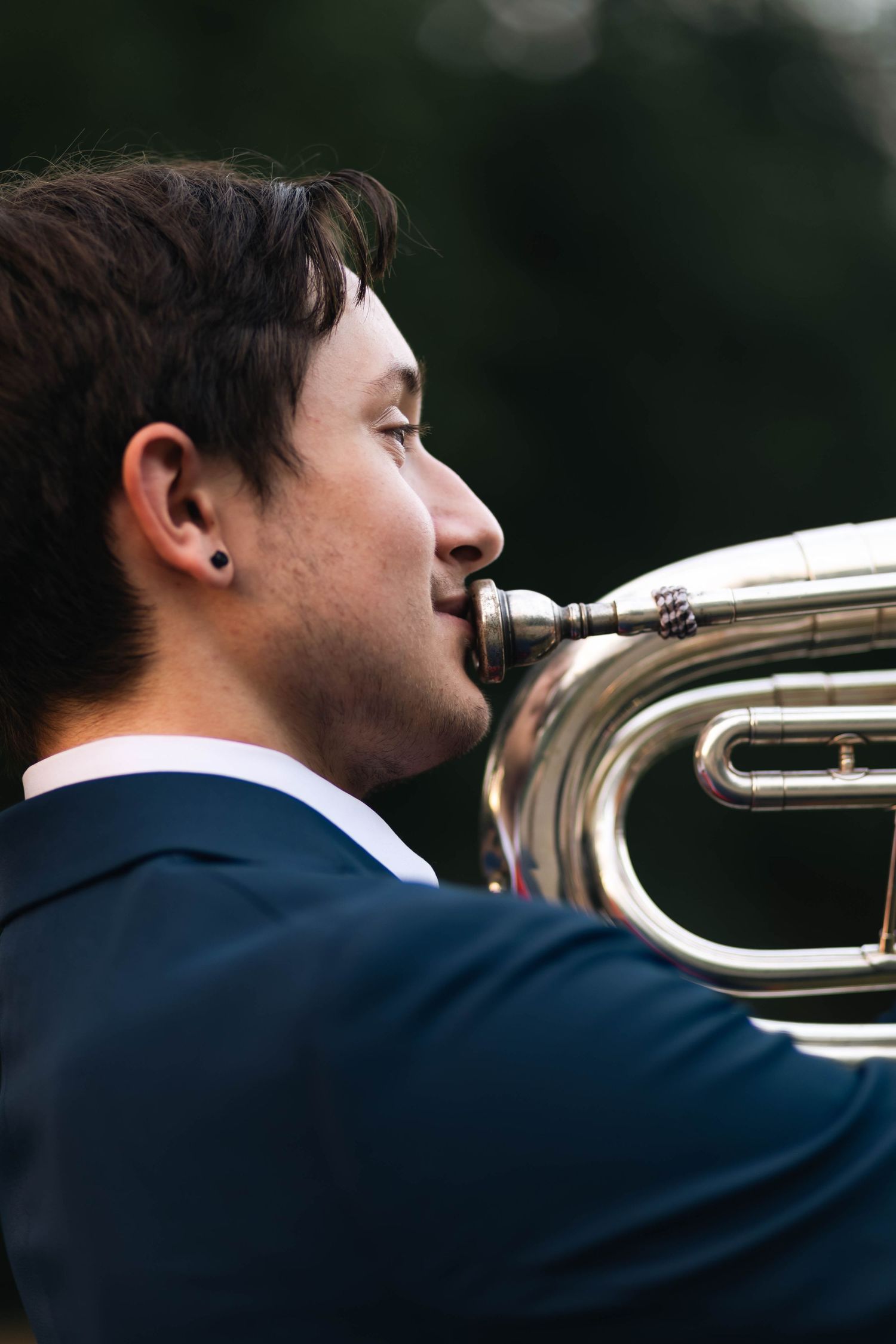 Young man in suit playing a euphonium brass instrument outdoors in profile view.