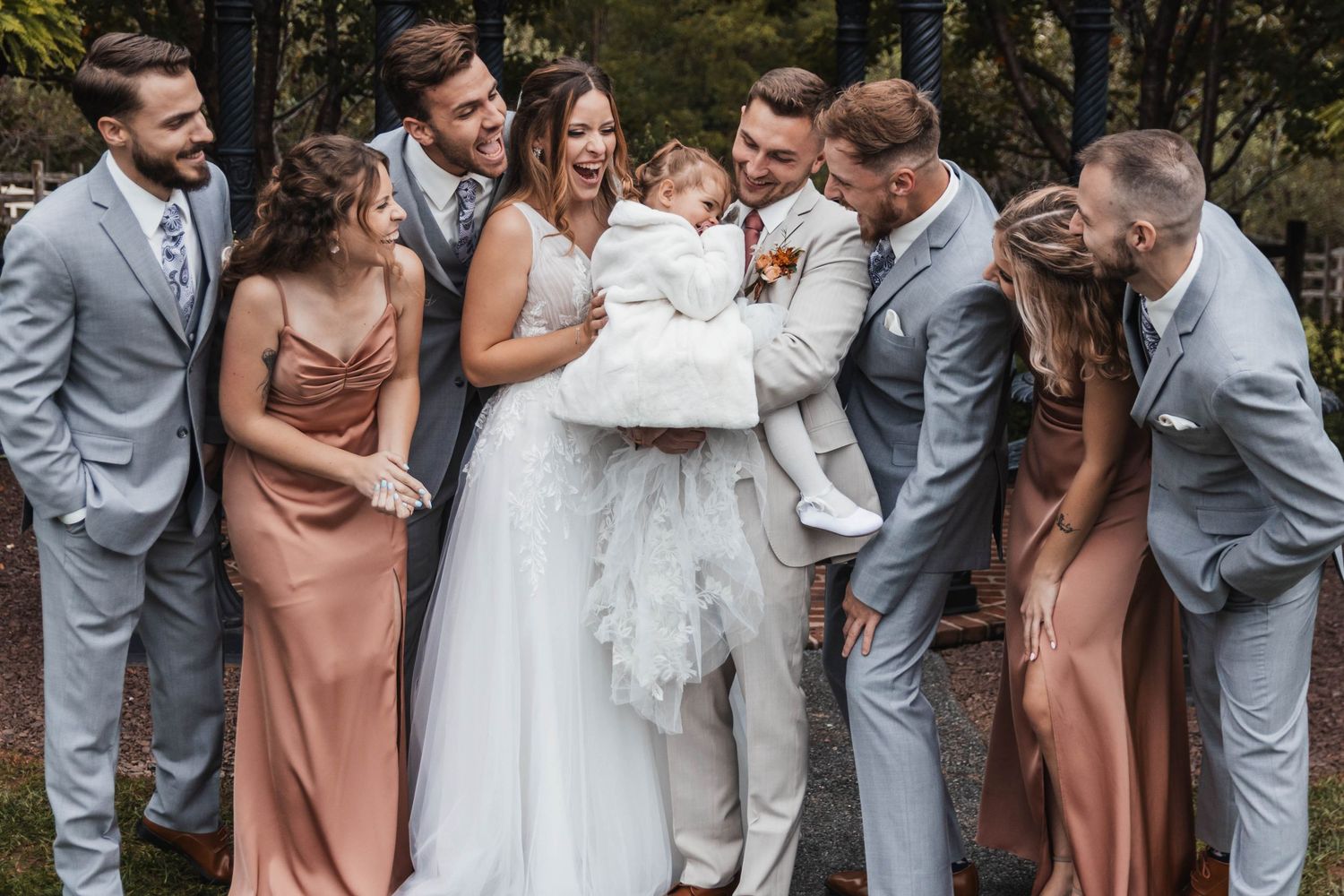 Bride and groom laugh joyfully surrounded by bridesmaids and groomsmen in dusty rose and gray attire.