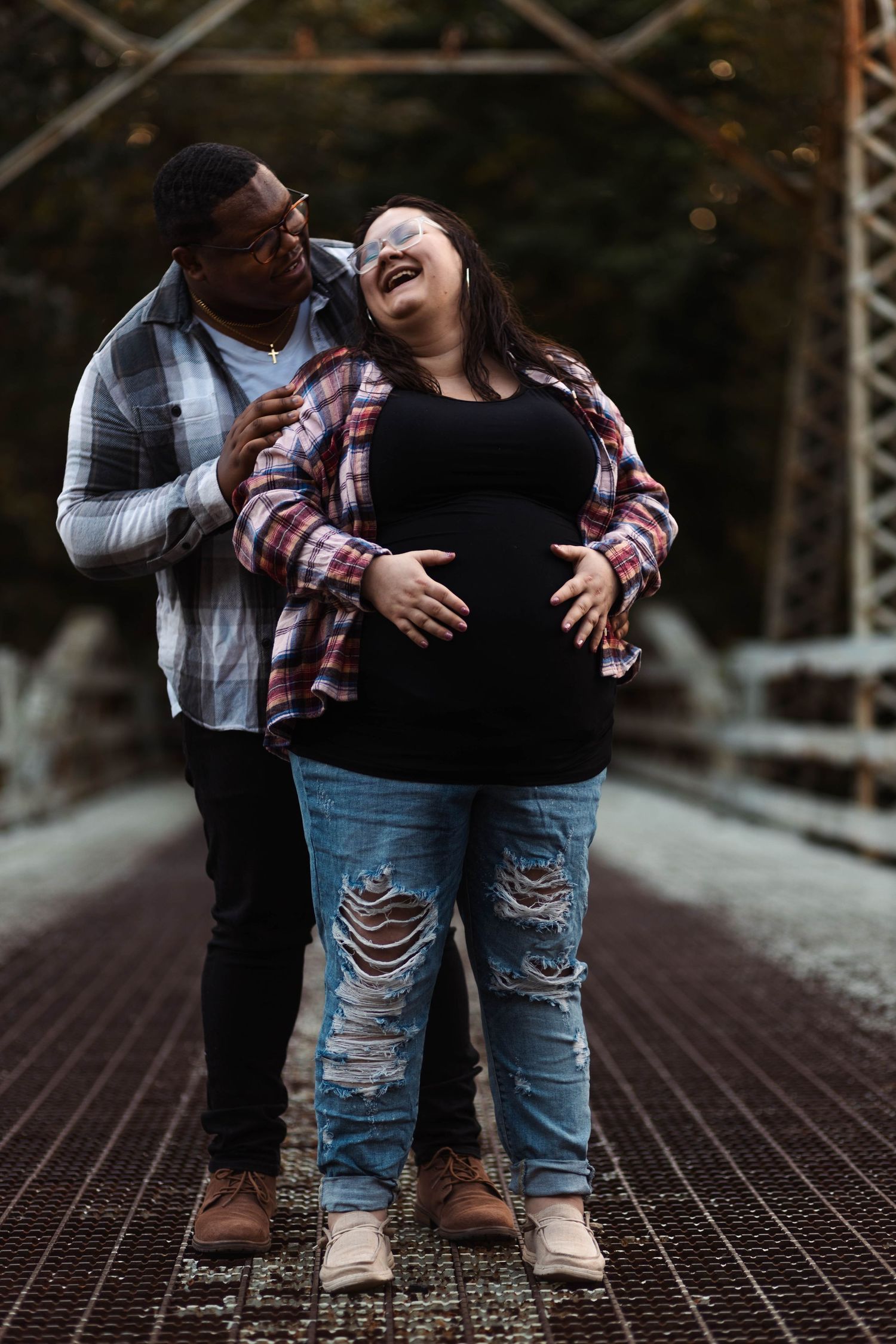 Laughing pregnant couple embraces on a bridge in autumn, man hugging woman from behind as she holds bump.