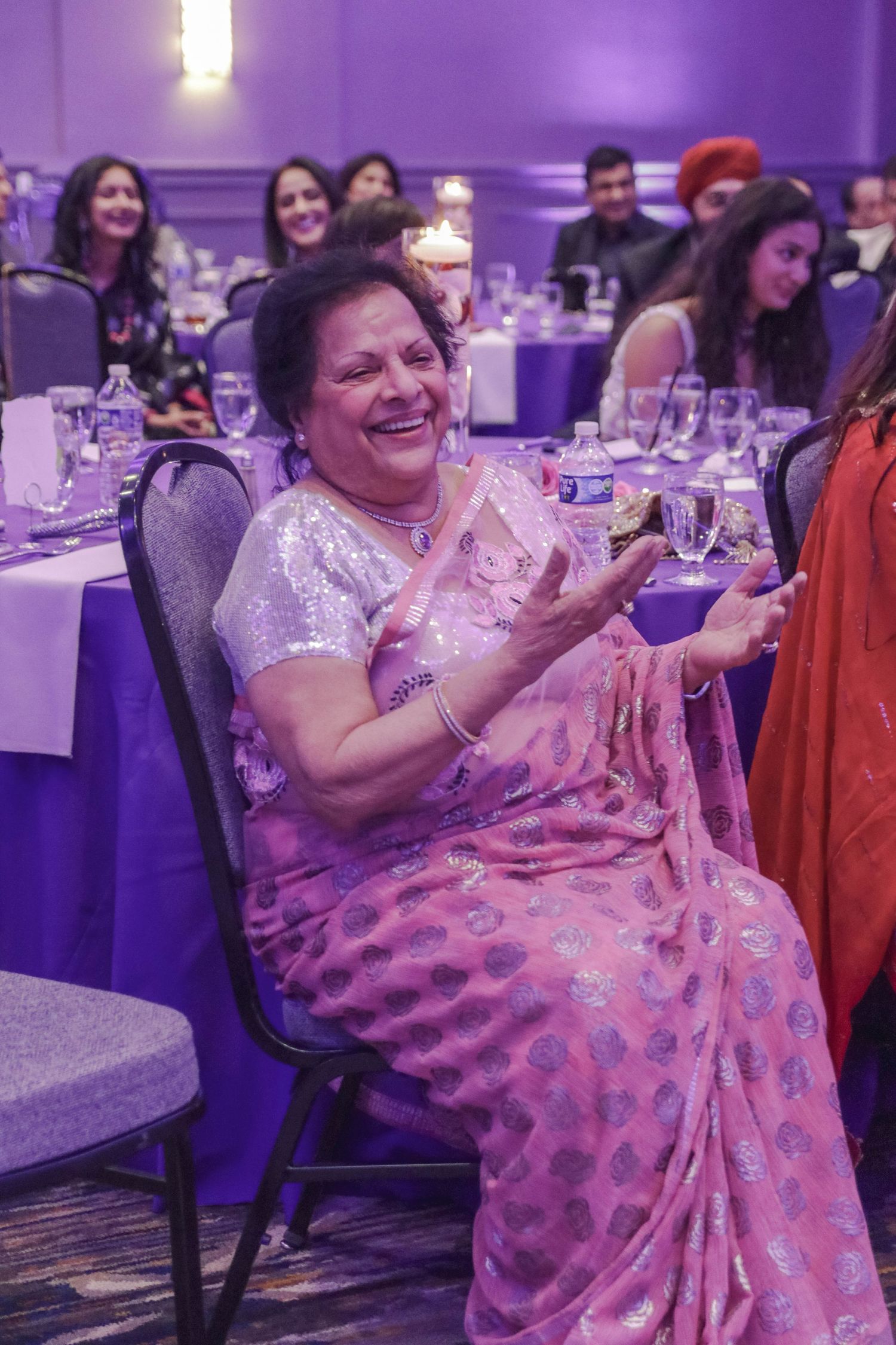 Smiling woman in pink and silver saree clapping at a festive Indian wedding banquet celebration.