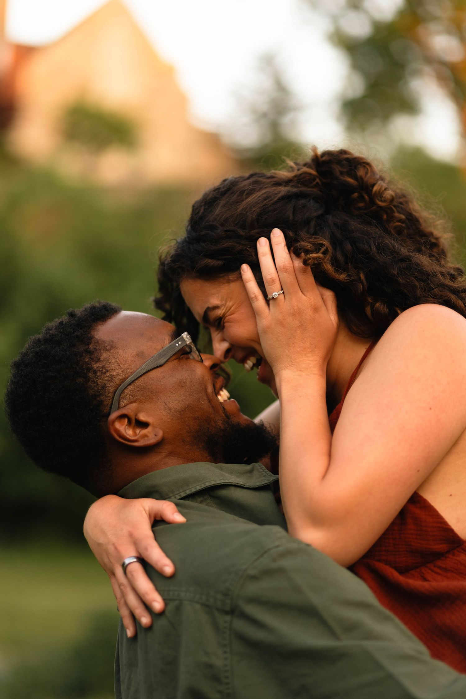 Joyful interracial couple sharing a romantic moment outdoors, woman showing engagement ring while being lifted by her partner.