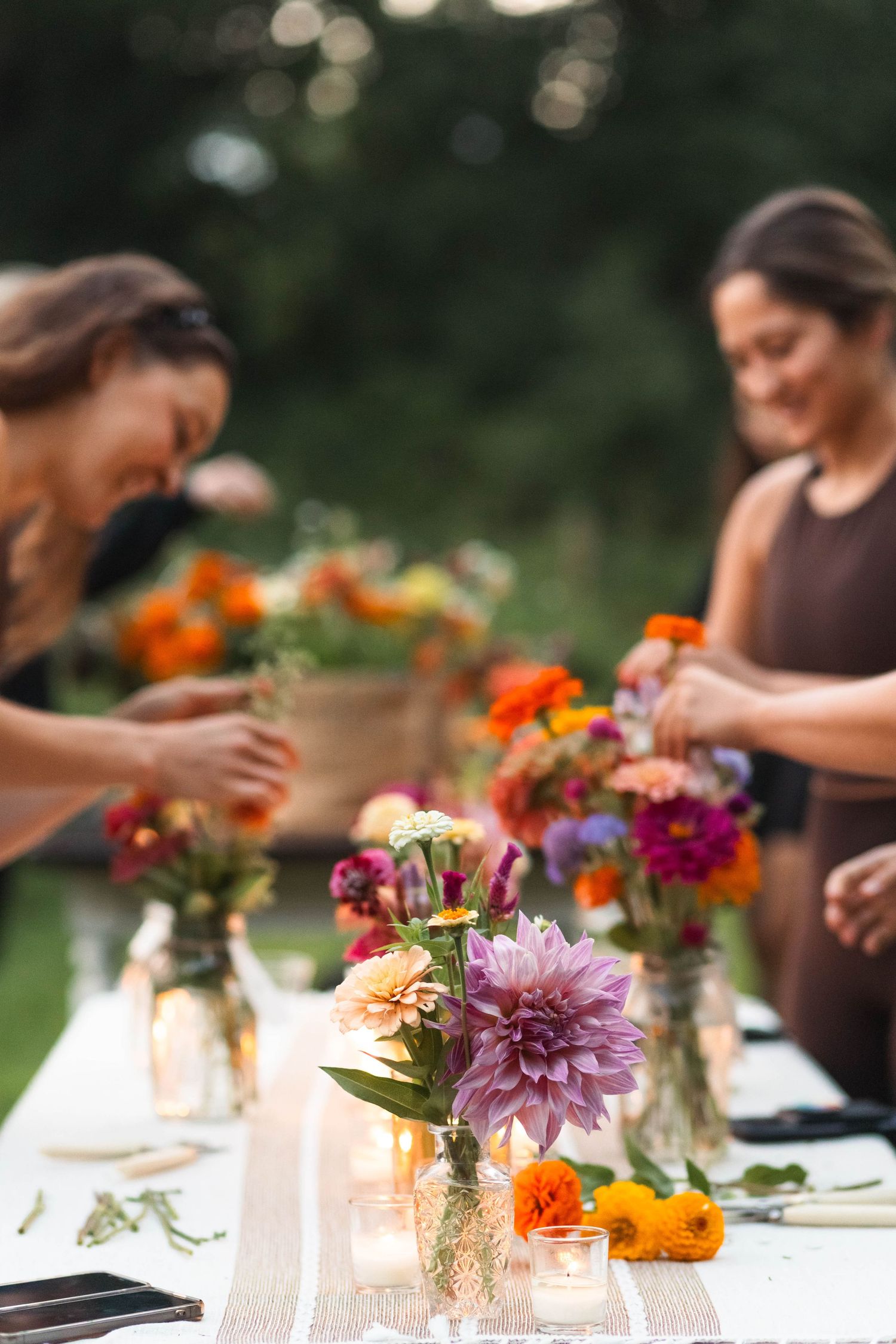 Two women arranging colorful flower bouquets in mason jars on a white outdoor dining table with candles.