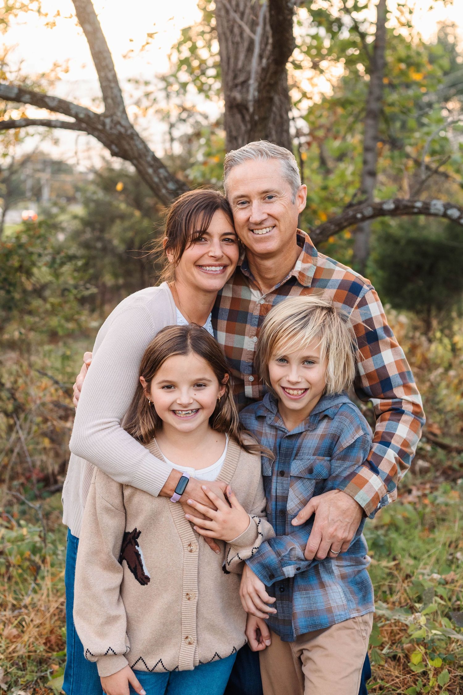 Happy family of four posing together outdoors among autumn trees in coordinating plaid and neutral outfits.