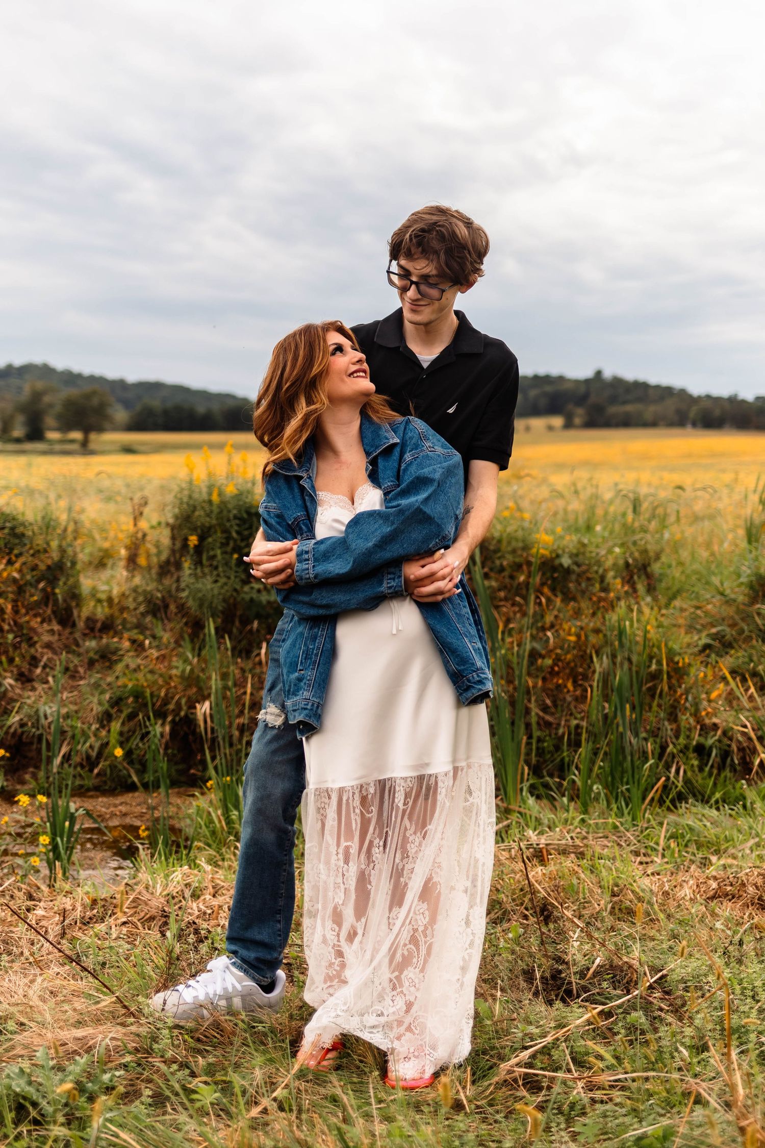 A young couple embraces in a golden field, woman in white lace dress and denim jacket, man in black shirt and jeans in Oley, PA