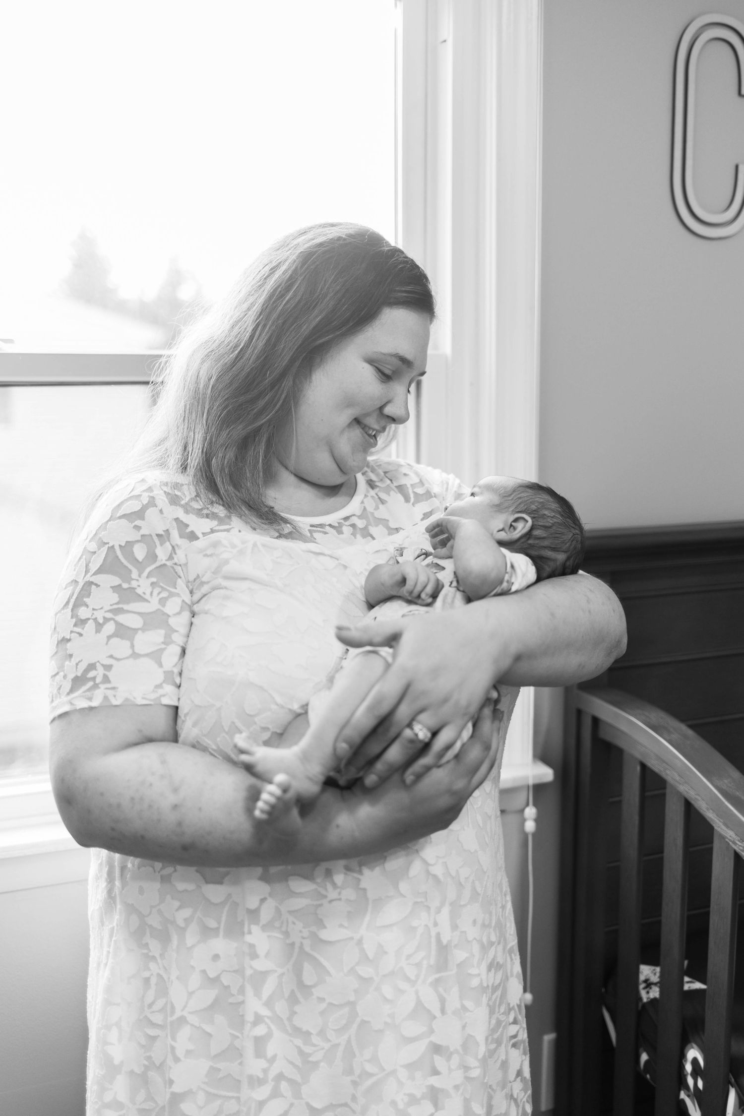 Black and white portrait of new mother in lace dress holding newborn by nursery window during an at home newborn session Philadelphia.