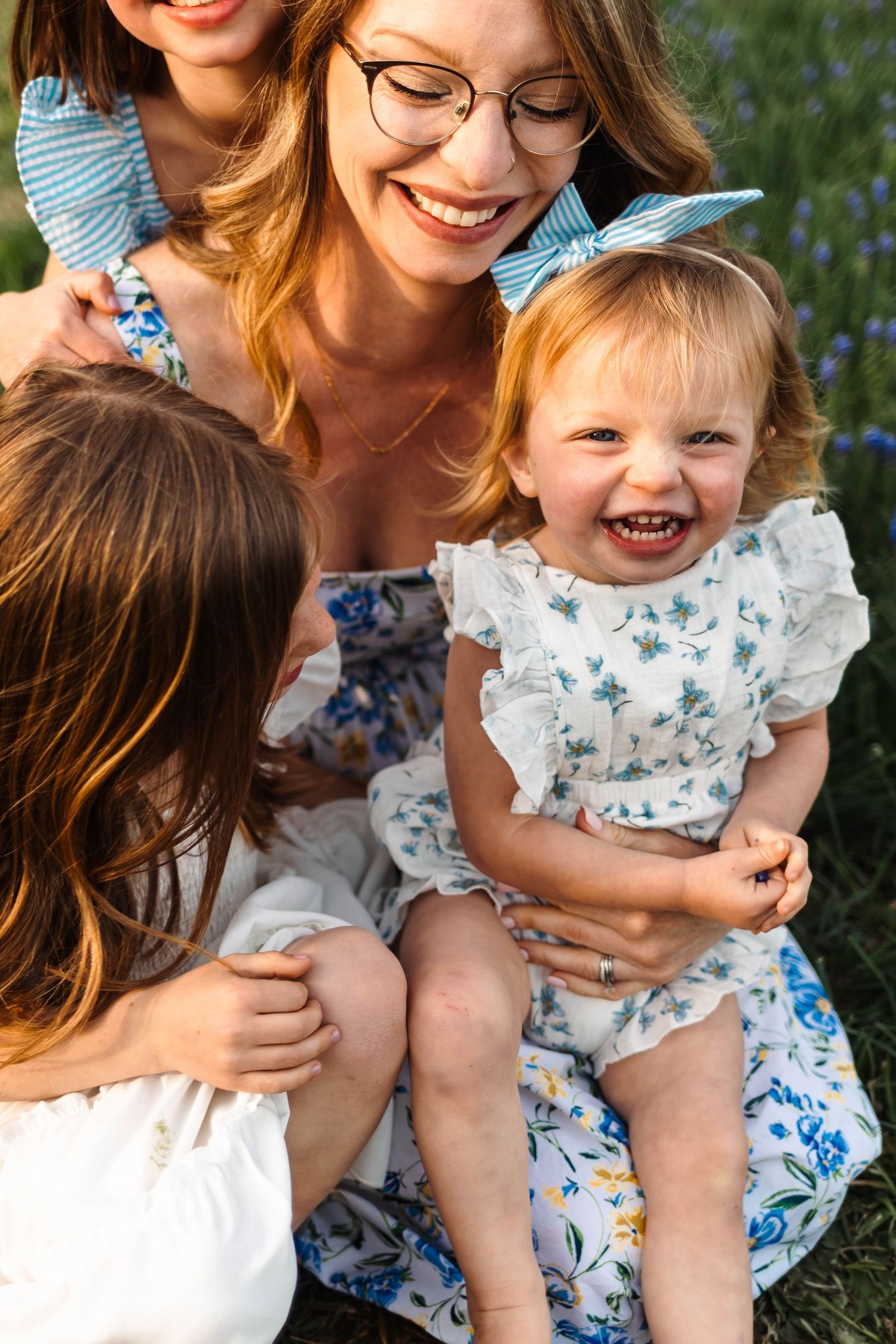 Smiling mother with glasses laughing with three young daughters in floral dresses in a garden.