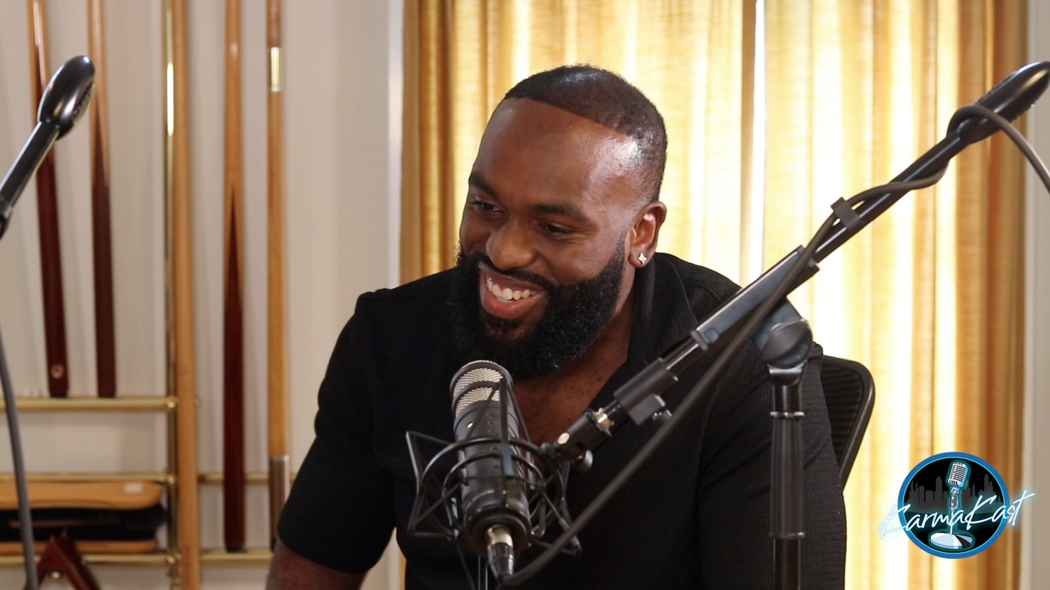 Smiling bearded Black man in black shirt speaking into a microphone at a podcast recording studio.