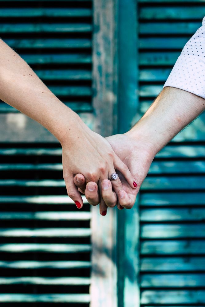 Close-up of an engaged couple holding hands showing diamond ring against teal wooden shutters.