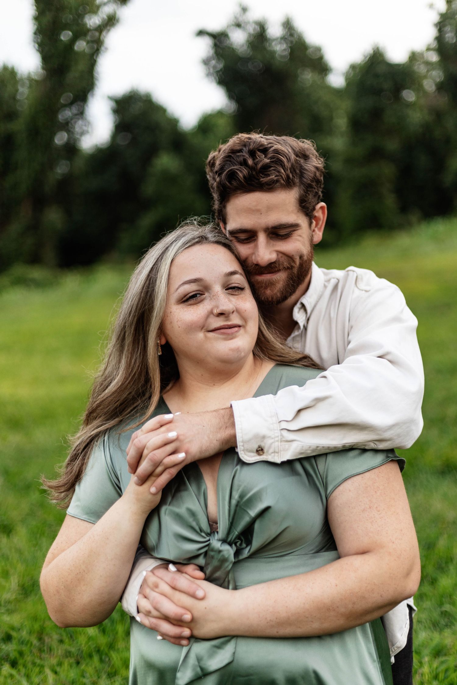 Bearded man embracing smiling woman in sage green dress in lush green outdoor field setting.