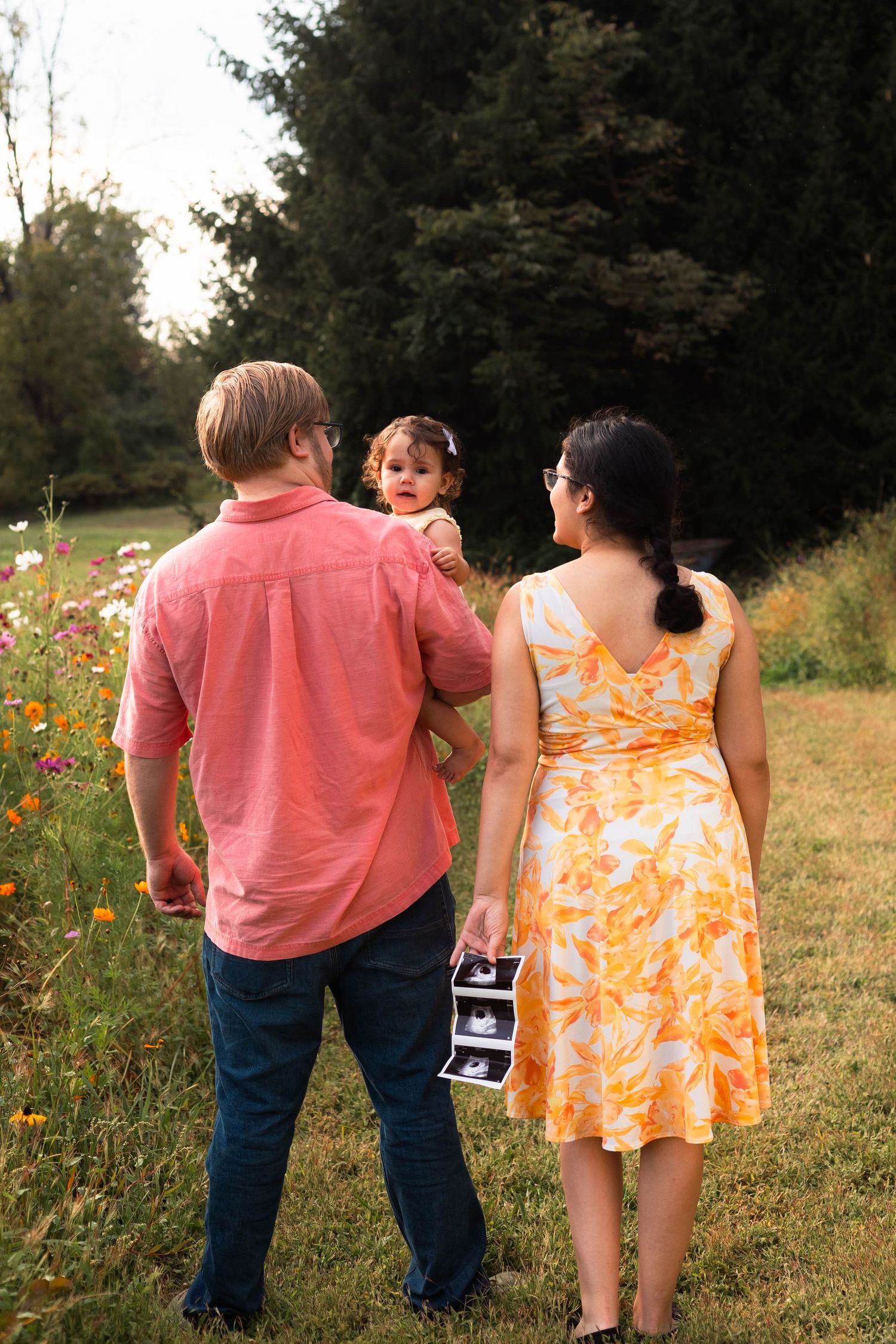 Family of three walking in flower field holding ultrasound photos from behind in Perkiomenville, PA.