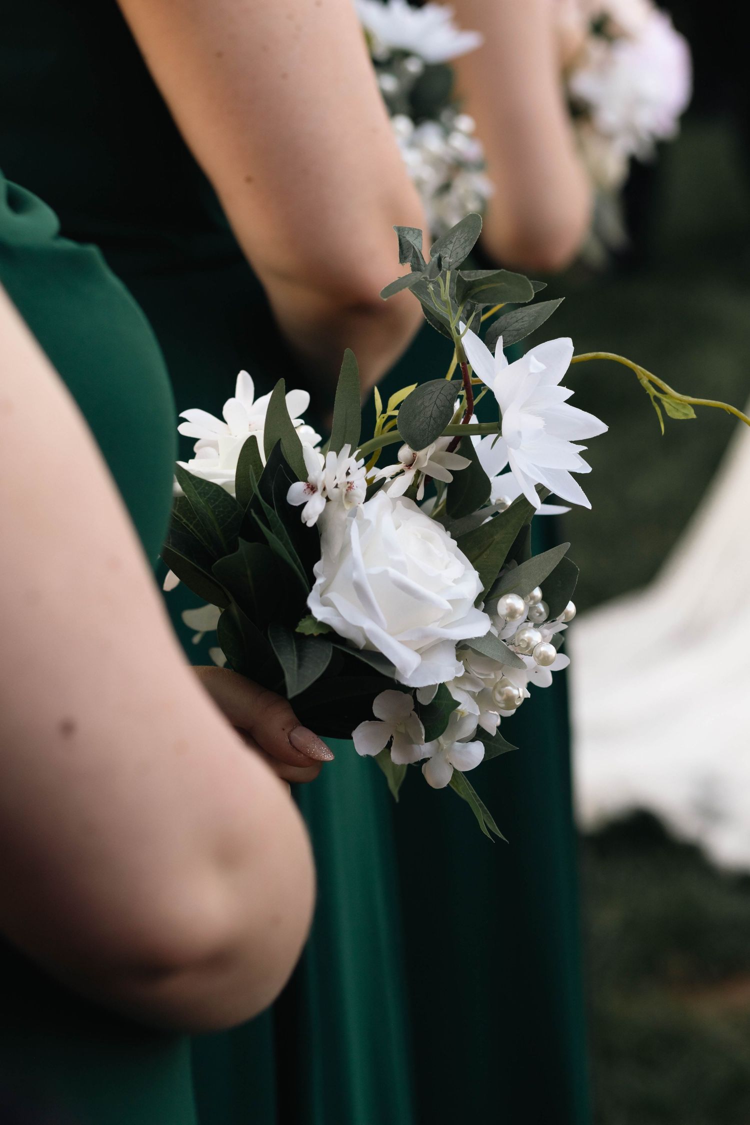 Bridesmaids in emerald green gowns holding elegant white rose and daisy bouquets with greenery at a wedding.