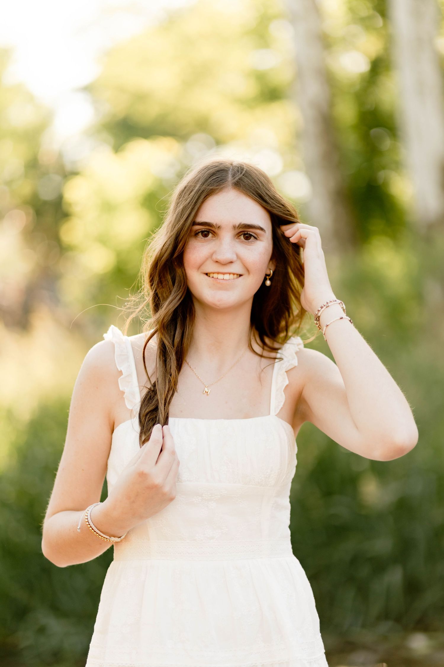 Smiling Boyertown High School Graduate in white ruffled dress touching her hair braid in a sunlit outdoor setting.