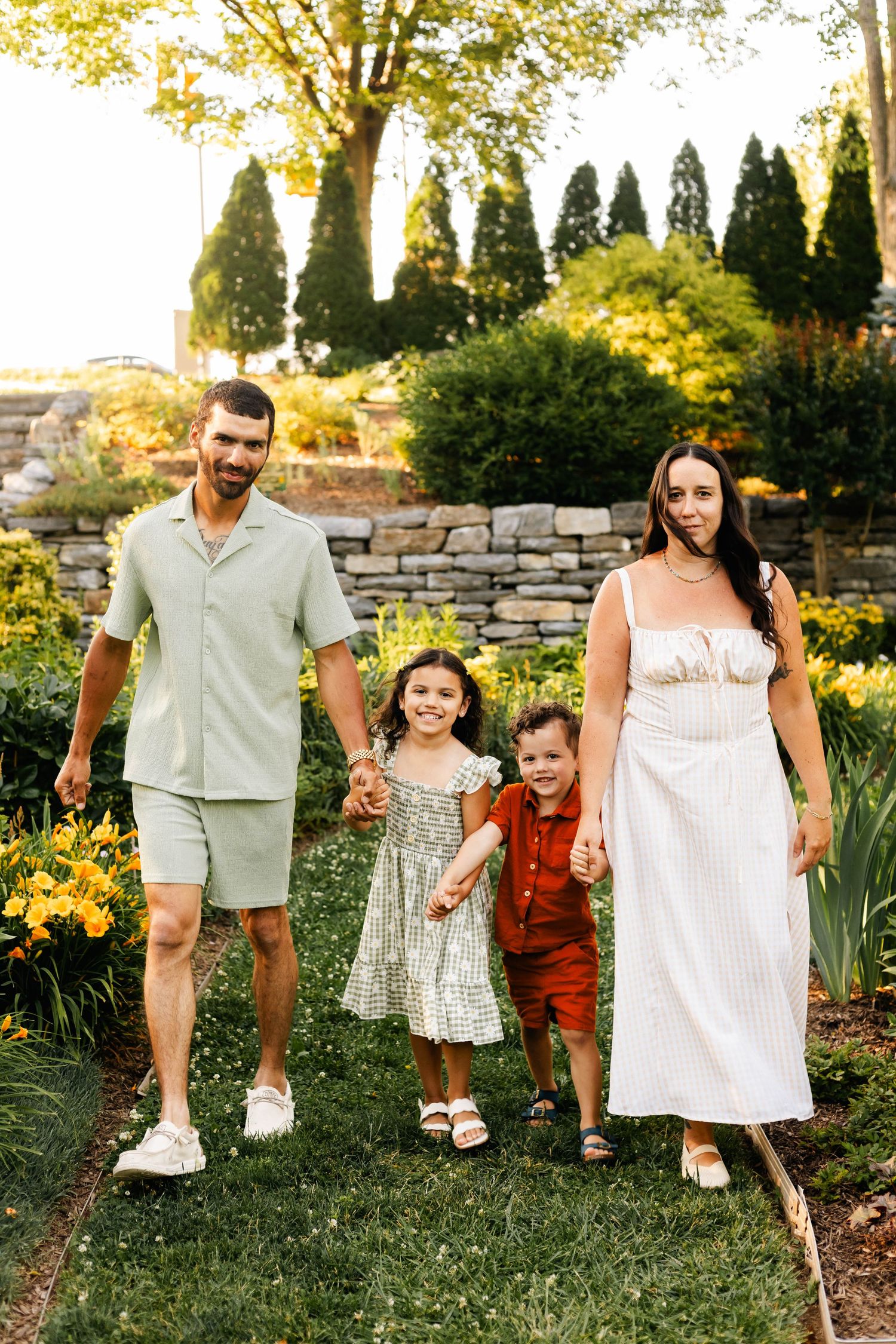 Family of four walking hand-in-hand through a lush garden with stone wall backdrop at sunset at Reading Museum Arboretum.