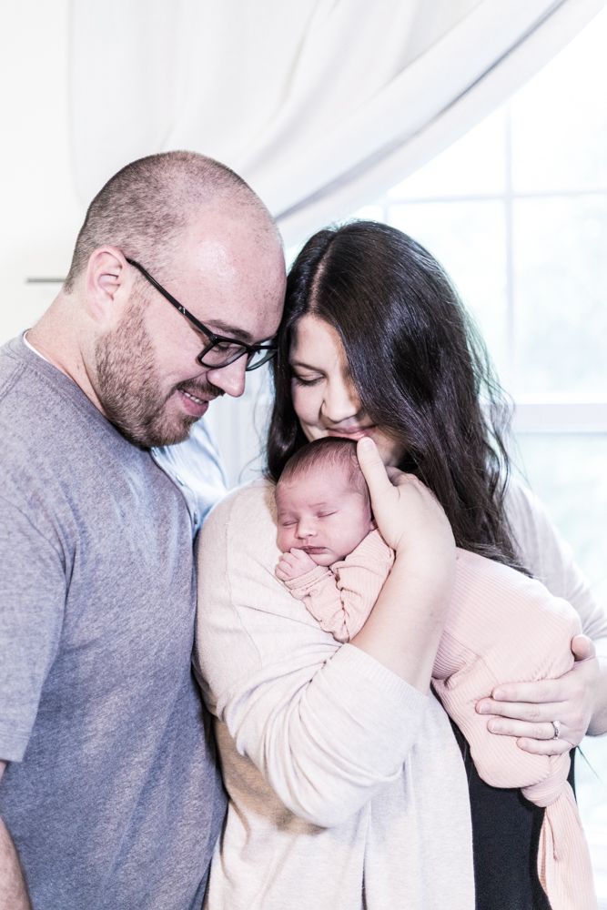 New parents tenderly holding sleeping newborn baby wrapped in pink blanket indoors.