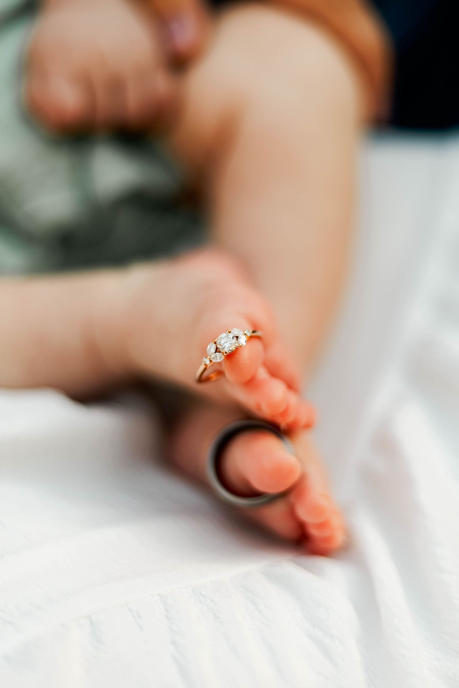 Close-up of a diamond engagement ring placed on tiny newborn baby toes on a white blanket.