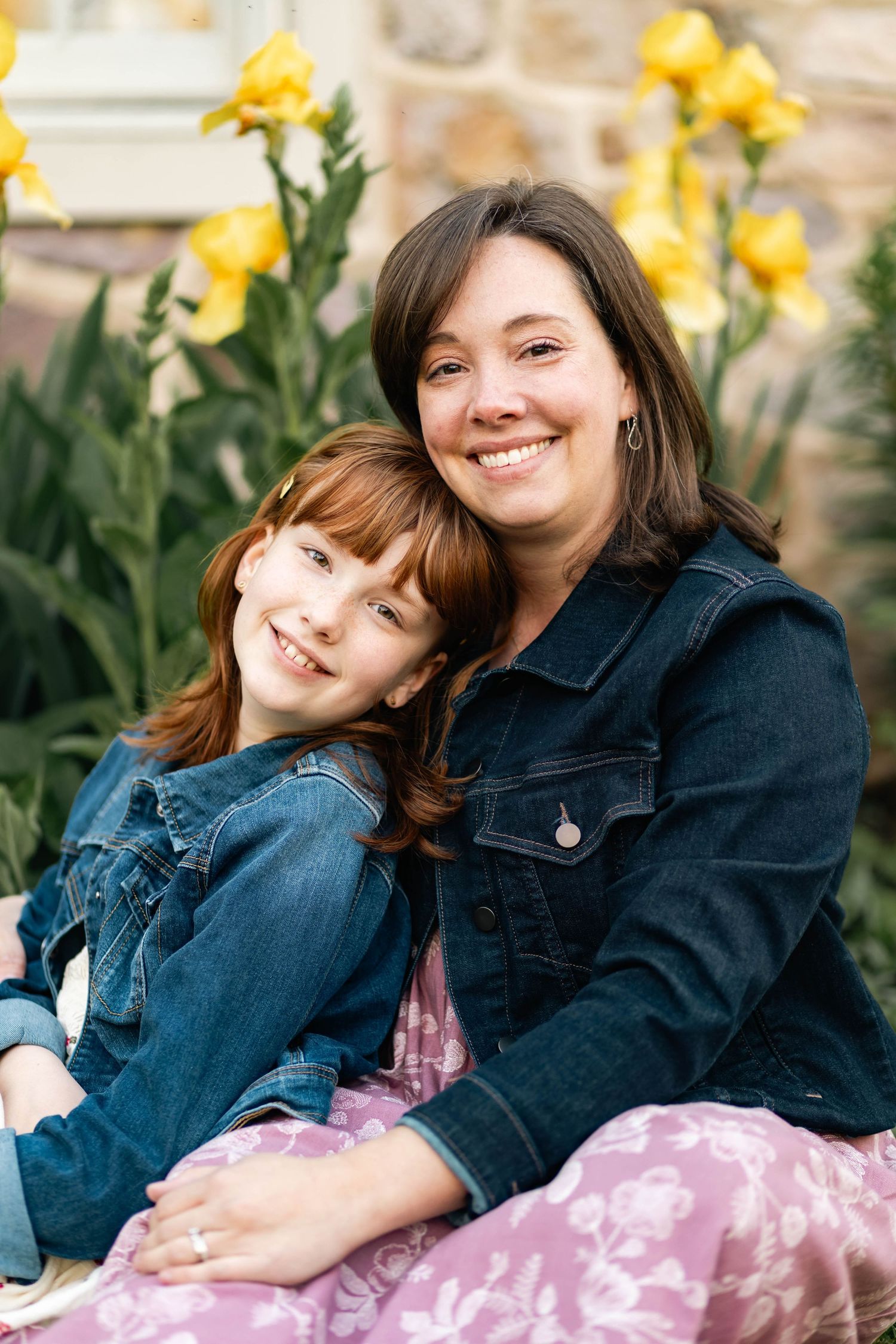 A smiling mother and red-haired daughter sit together in front of bright yellow flowers in an outdoor garden in Elverson, PA.