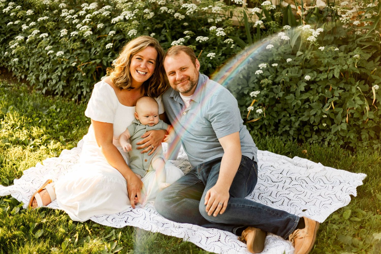 Smiling family of three sits on a white blanket in a sunny garden with their baby in Elverson, PA.