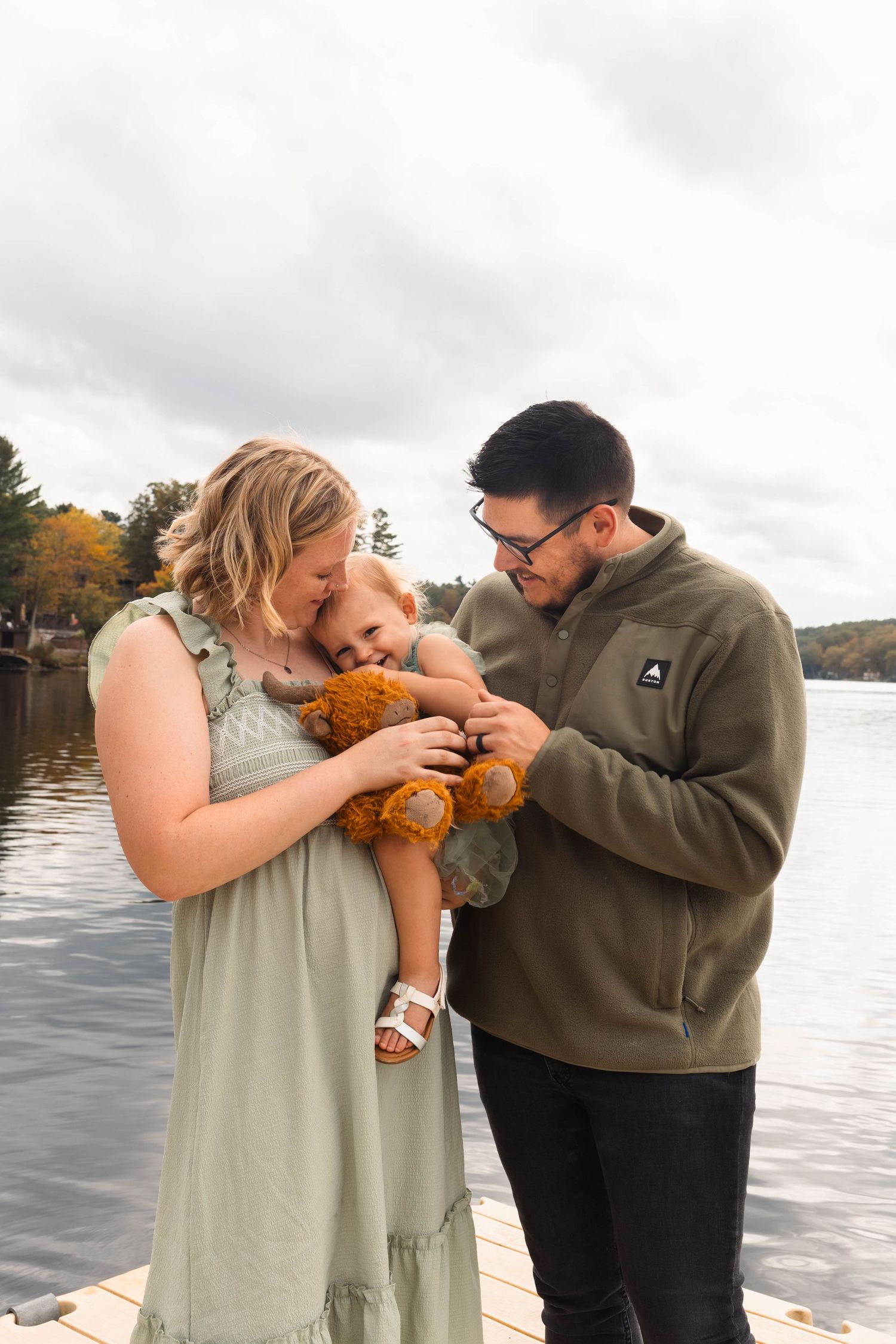 A couple shares a tender moment with their baby holding a teddy bear on a dock by a lake in autumn in the Poconos.