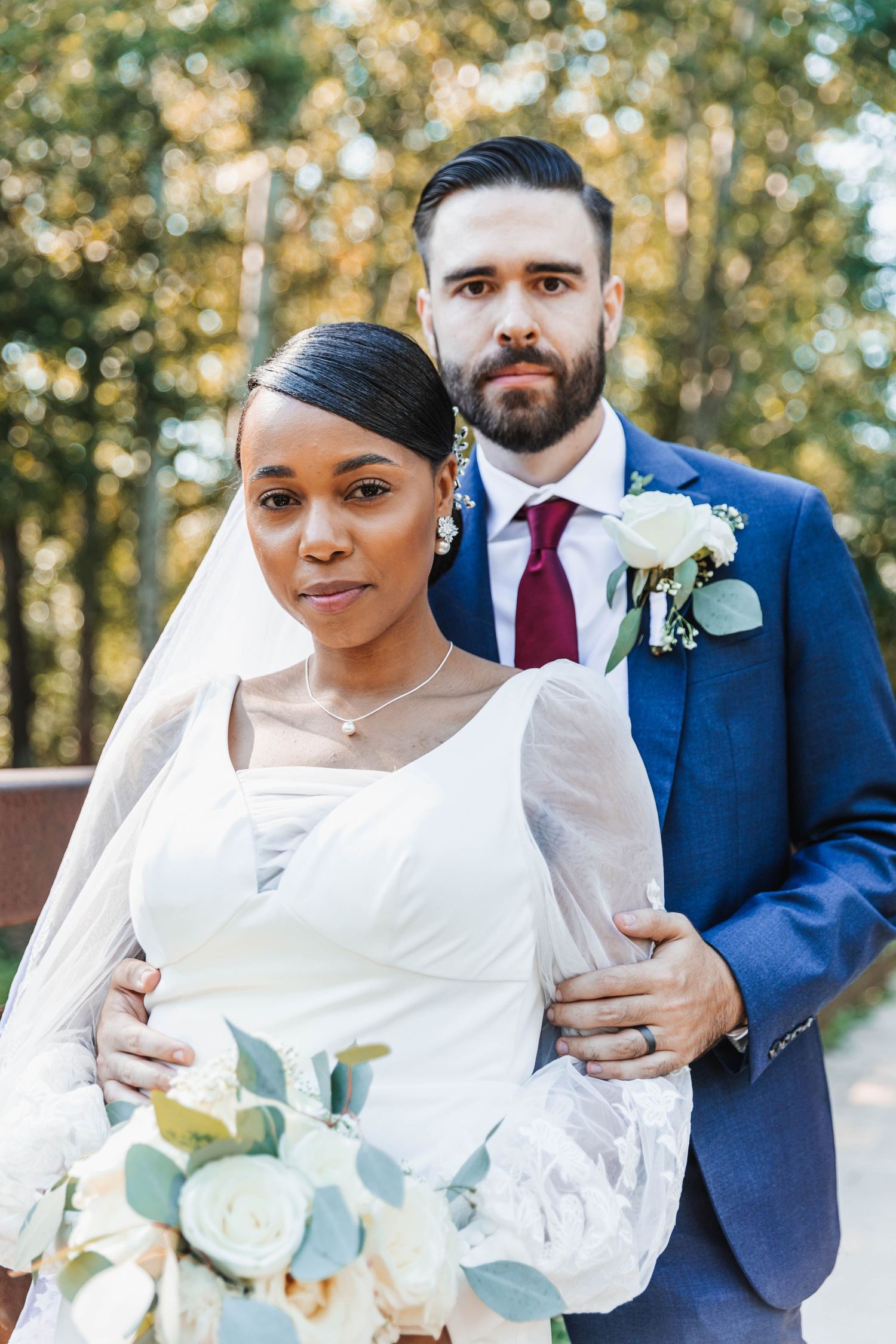 Interracial bride and groom posing in autumn forest, groom in navy suit with red tie.