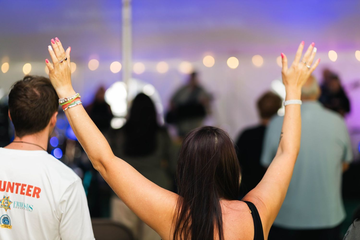 Two people raising hands in worship at an outdoor tent revival event in Boyertown, PA with bokeh string lights background.