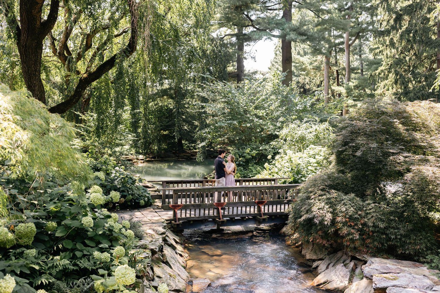 Couple standing on a wooden bridge surrounded by lush green gardens and tall trees in a serene outdoor setting.