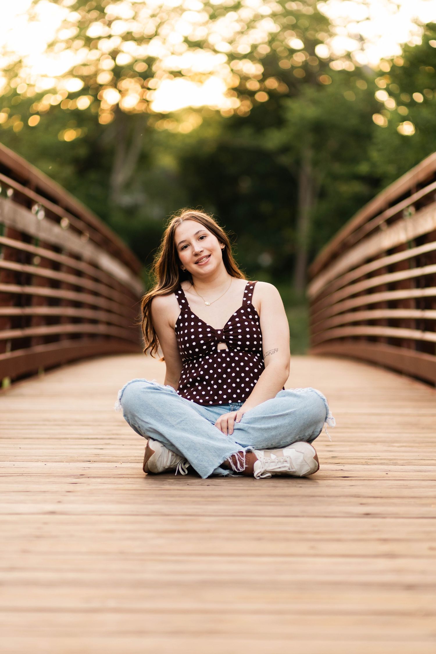 Boyertown Senior Session in polka dot top and jeans sitting cross-legged on a wooden bridge at sunset.