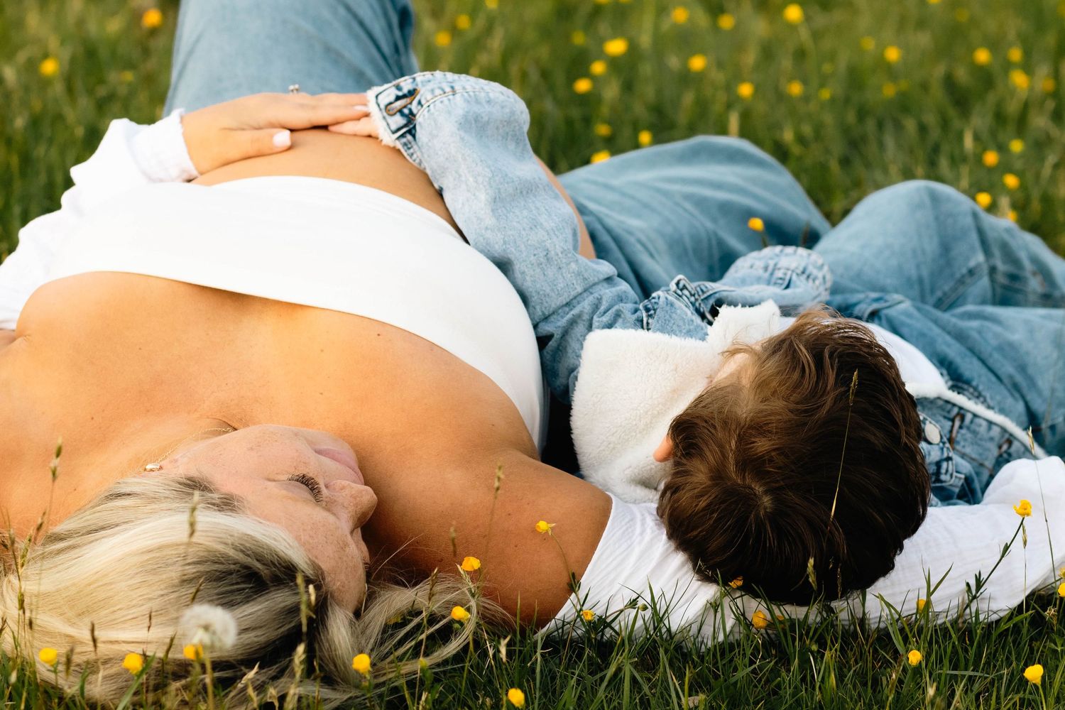 Couple lying in green grass field with yellow flowers, man resting head near pregnant womans belly at Hopewell Furnace in Elverson, PA.
