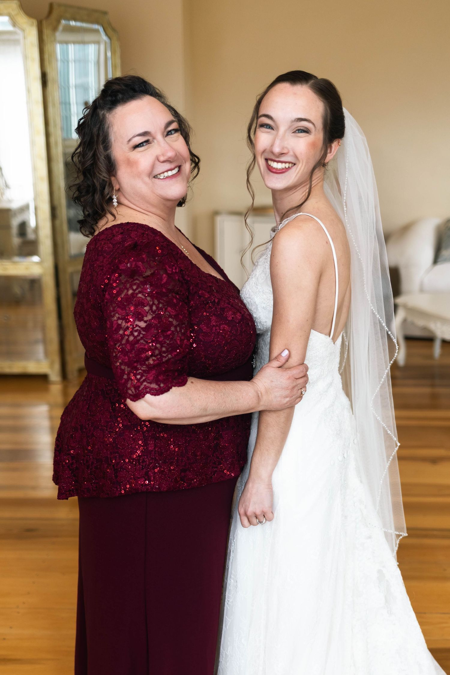 Smiling bride in white lace gown with veil poses with mother in burgundy lace dress.
