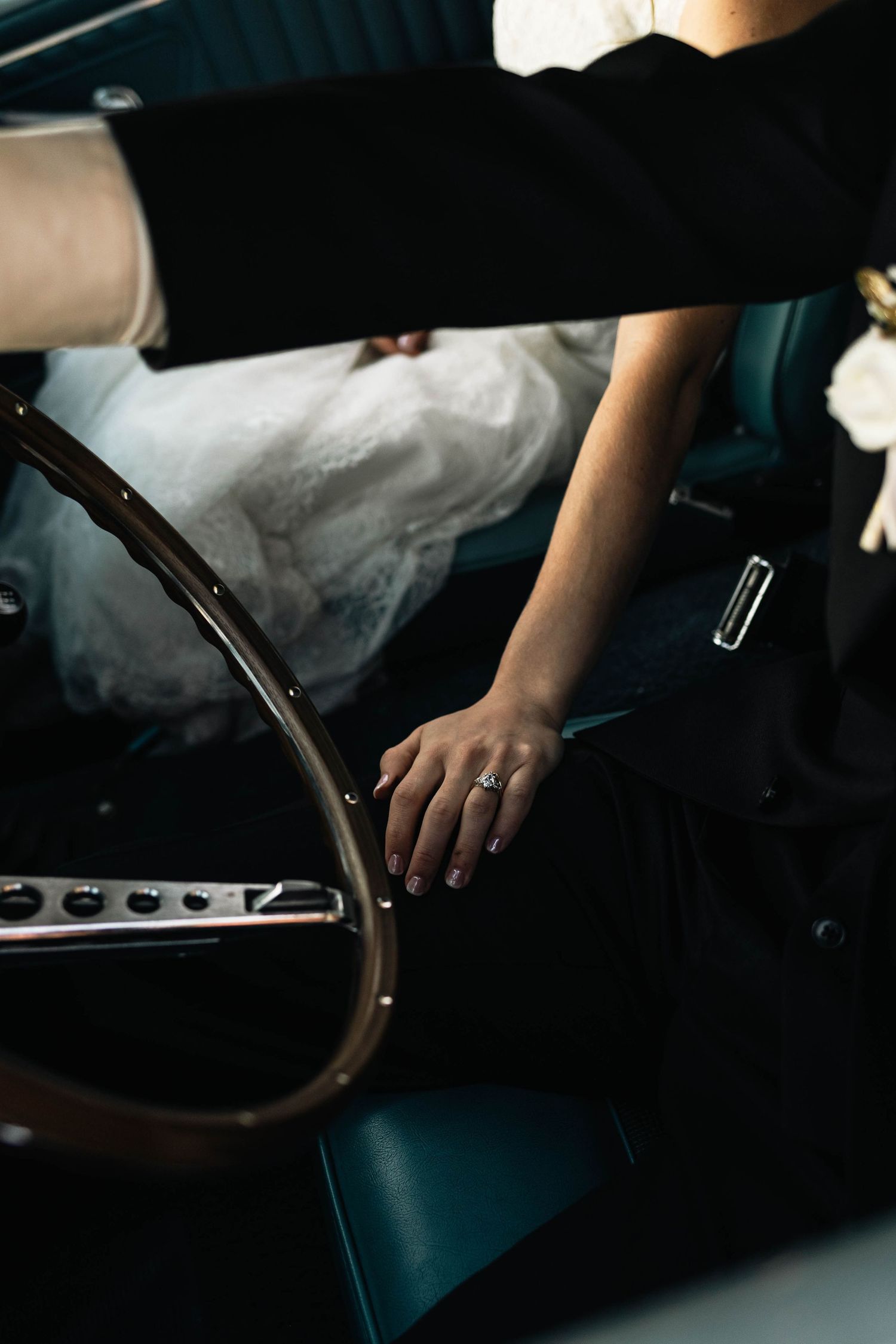Close-up of bride's hand with engagement ring resting on groom's leg in a vintage car during wedding day.