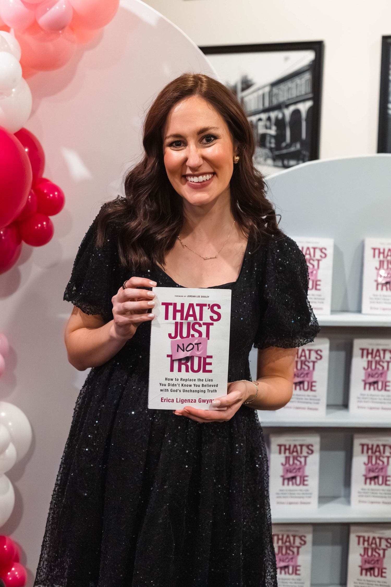 A smiling woman holds up her book That's Just Not True at a colorful book launch party with balloons at Brakeman's Cafe in Boyertown.